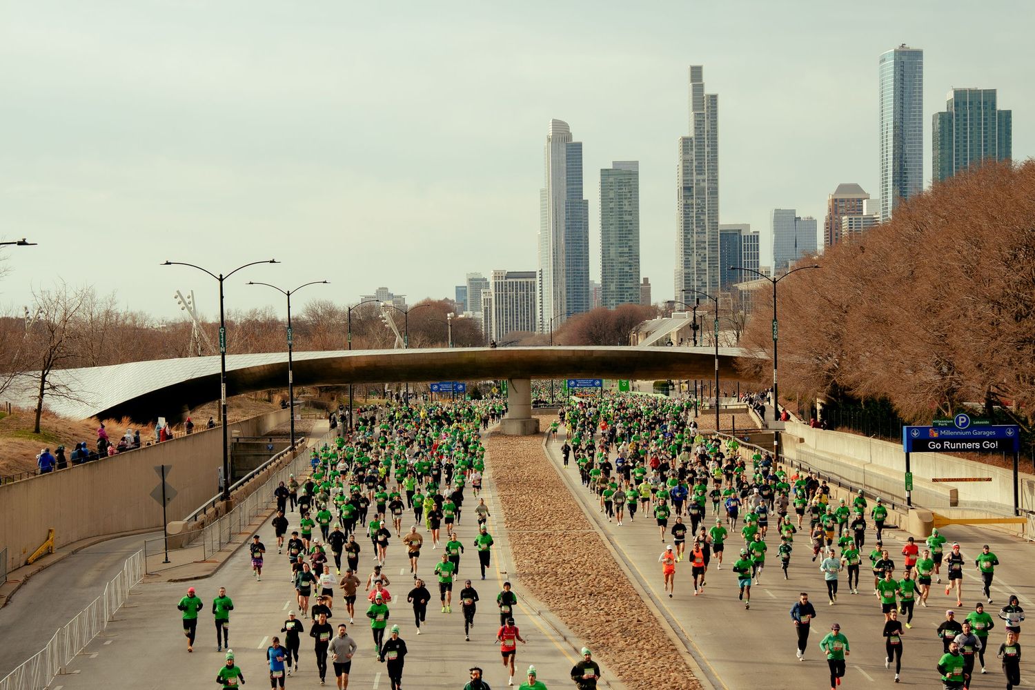 Large crowd of runners in green attire racing through Chicago streets with city skyline backdrop during St. Patrick's Day race.