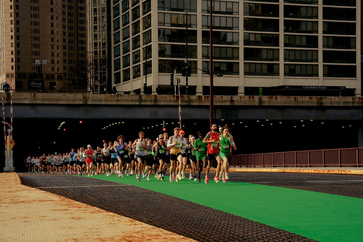 Runners compete in a city marathon race along a green-painted urban course with skyscrapers in the background.