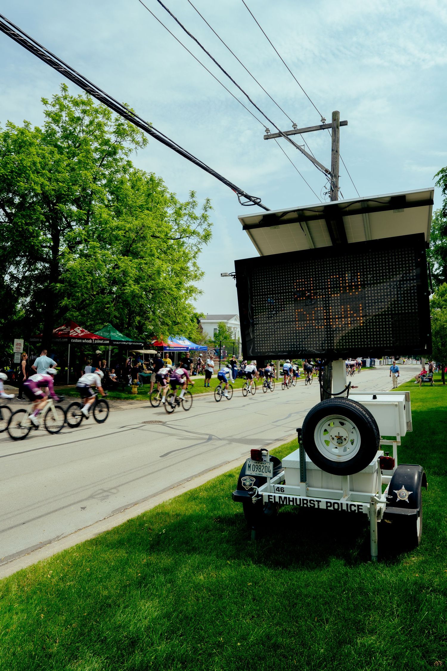 Digital display board on trailer beside bike path with cyclists passing by on sunny day.