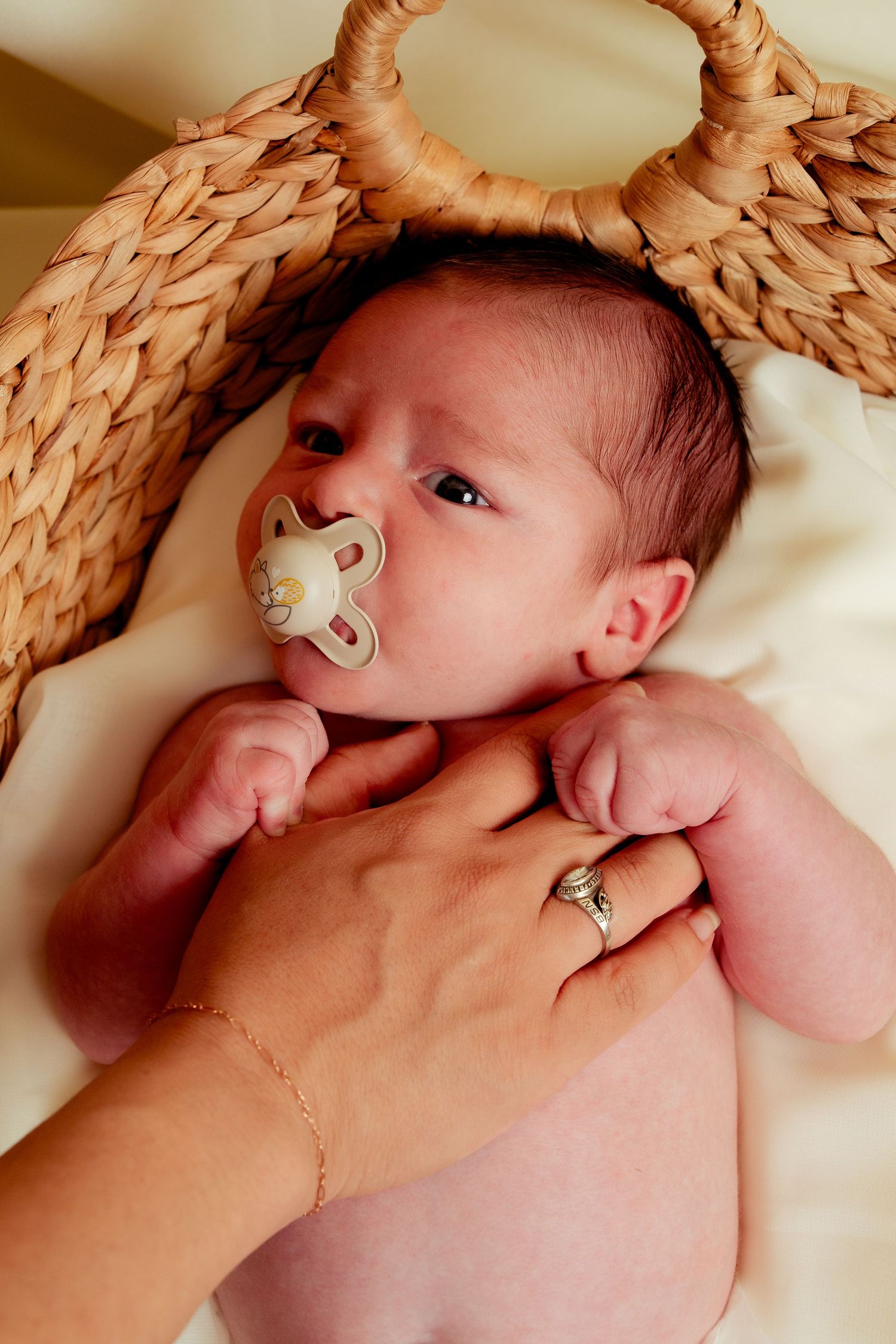 Newborn baby with pacifier lying in wicker basket, parent's hand gently resting on chest, wearing silver ring.