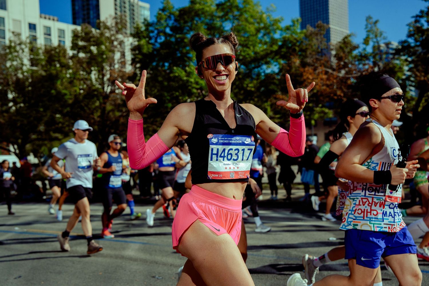 Marathon runner in pink shorts showing peace signs while running through city streets.