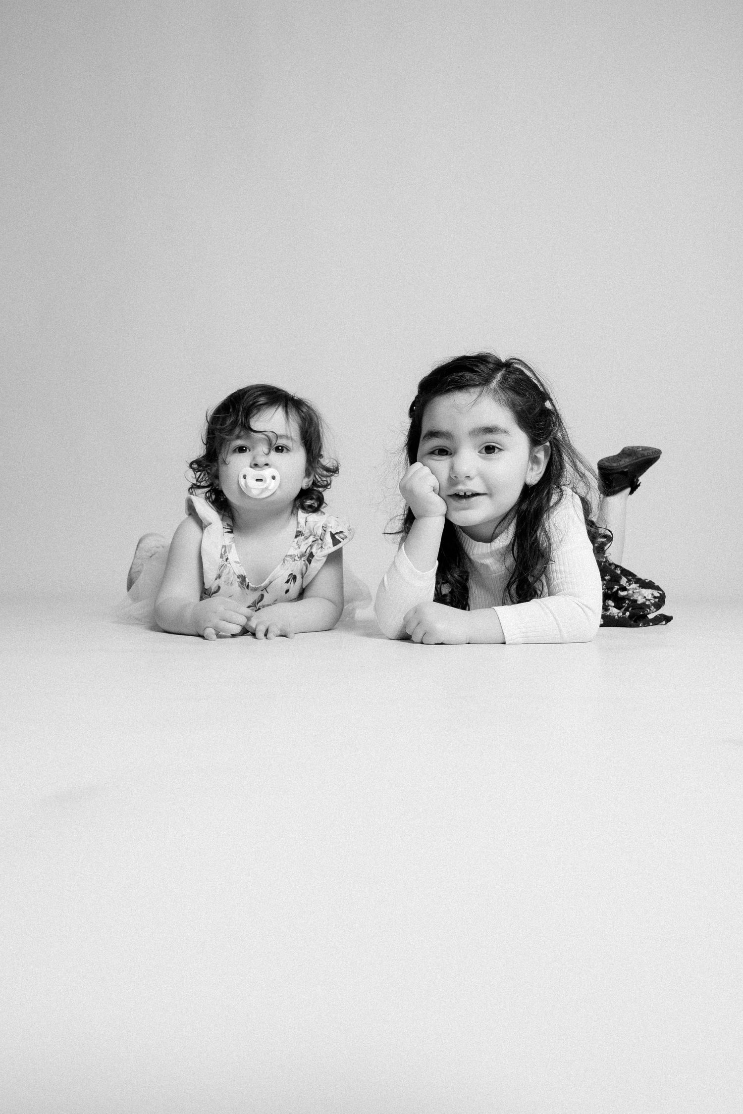 Two young girls lying on floor in black and white studio portrait, one with pacifier, posing together.