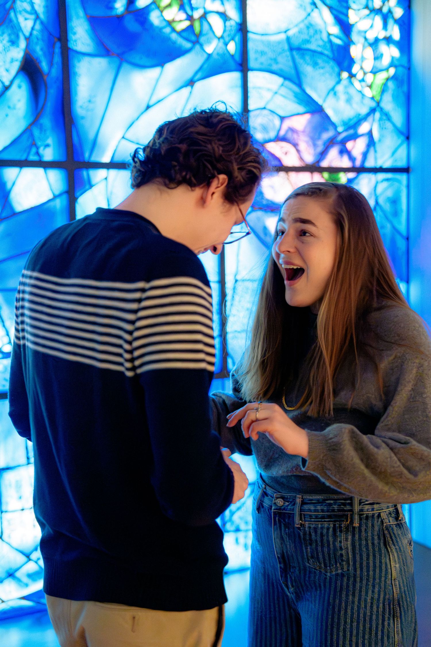 A couple shares a joyful moment in front of vibrant blue stained glass windows.