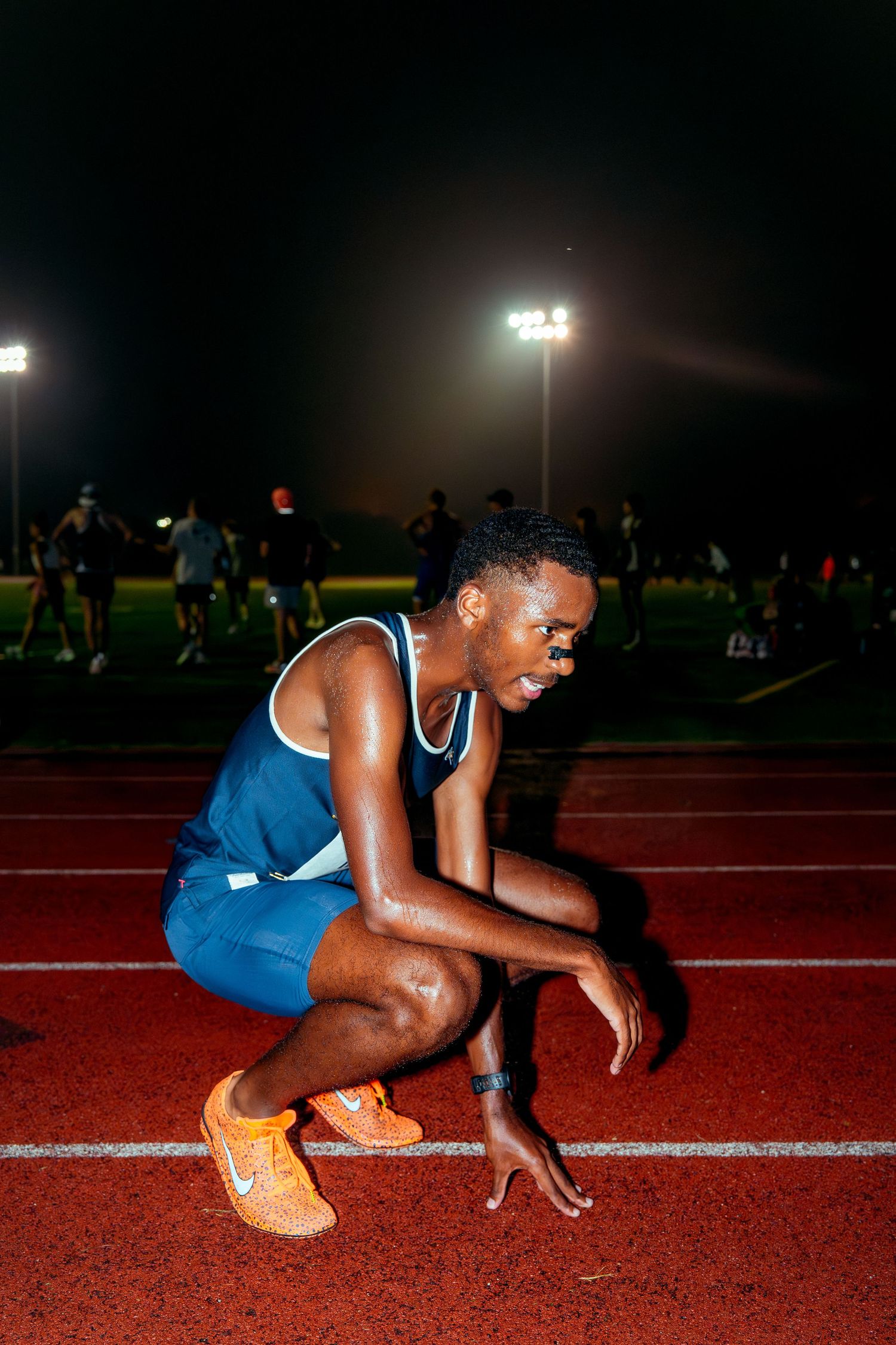 Athlete in blue uniform crouching at starting position on red track under stadium lights.