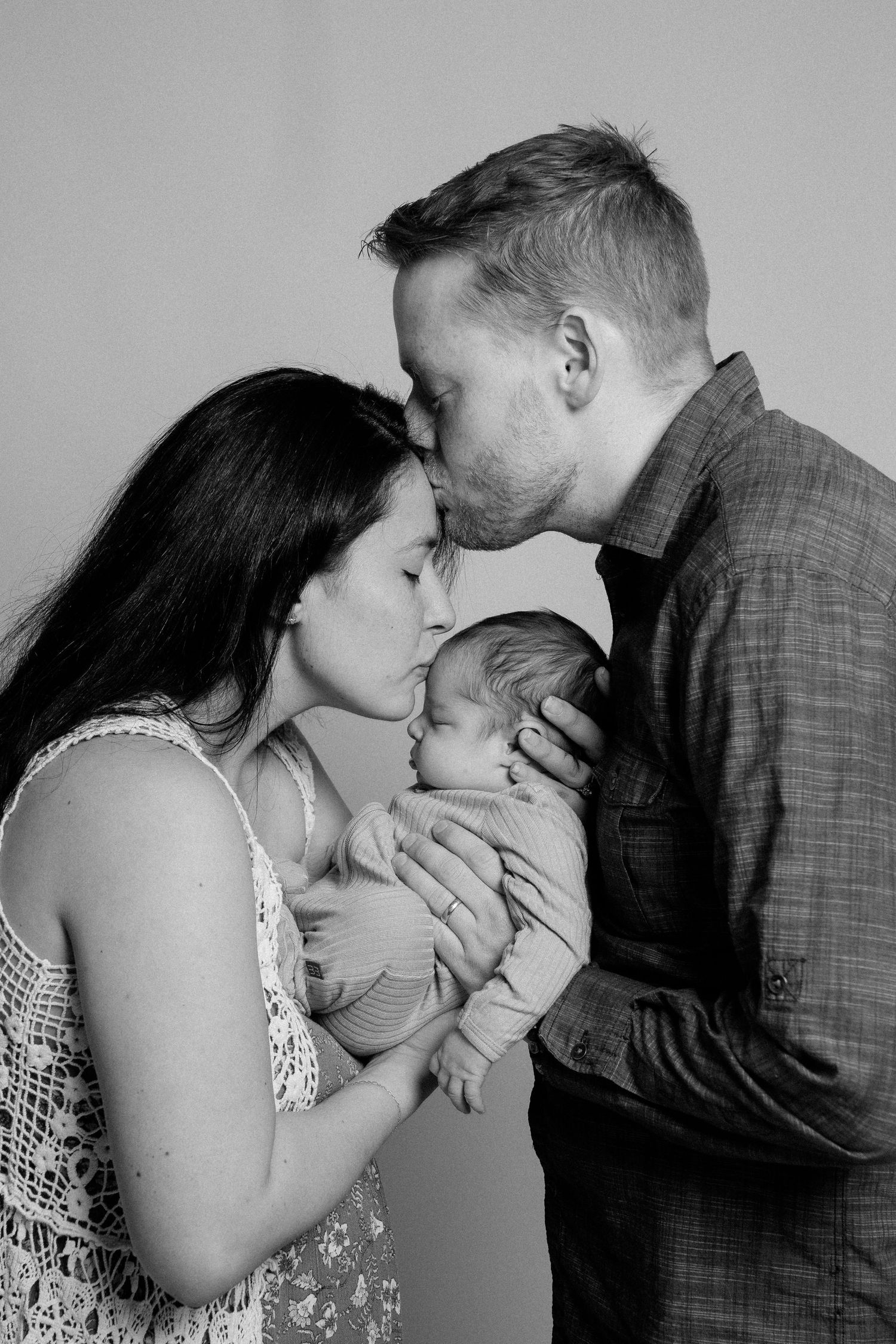 Black and white photo of parents kissing their newborn baby, a tender family portrait capturing a precious moment.
