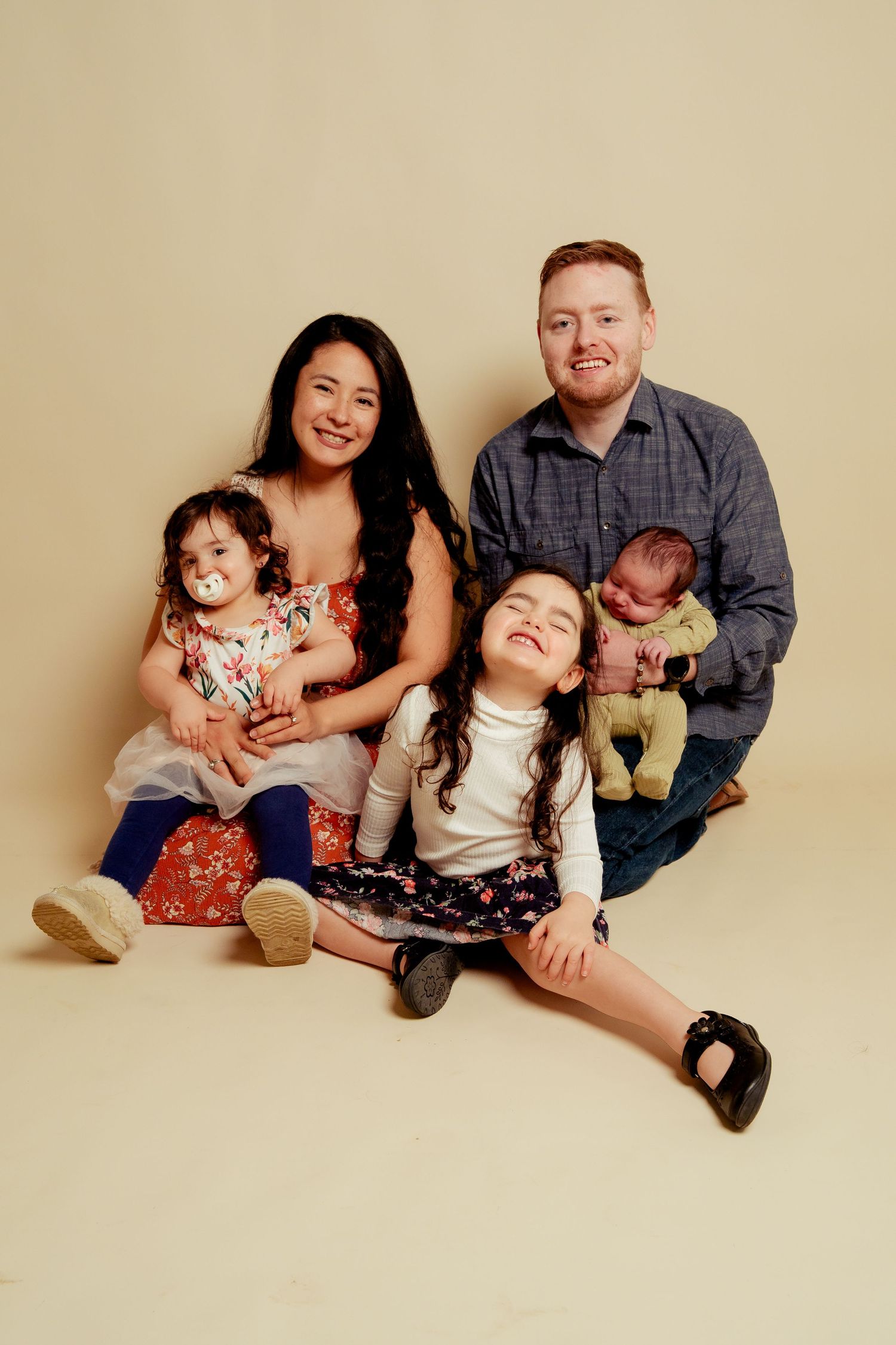 A happy family of five poses for a studio portrait, with parents holding three young children against a beige background.