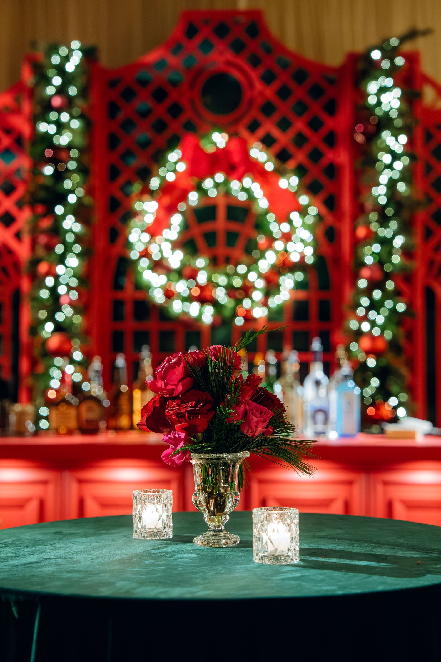 Red trellis backdrop with white Christmas lights and wreath, featuring roses in crystal vase on table.
