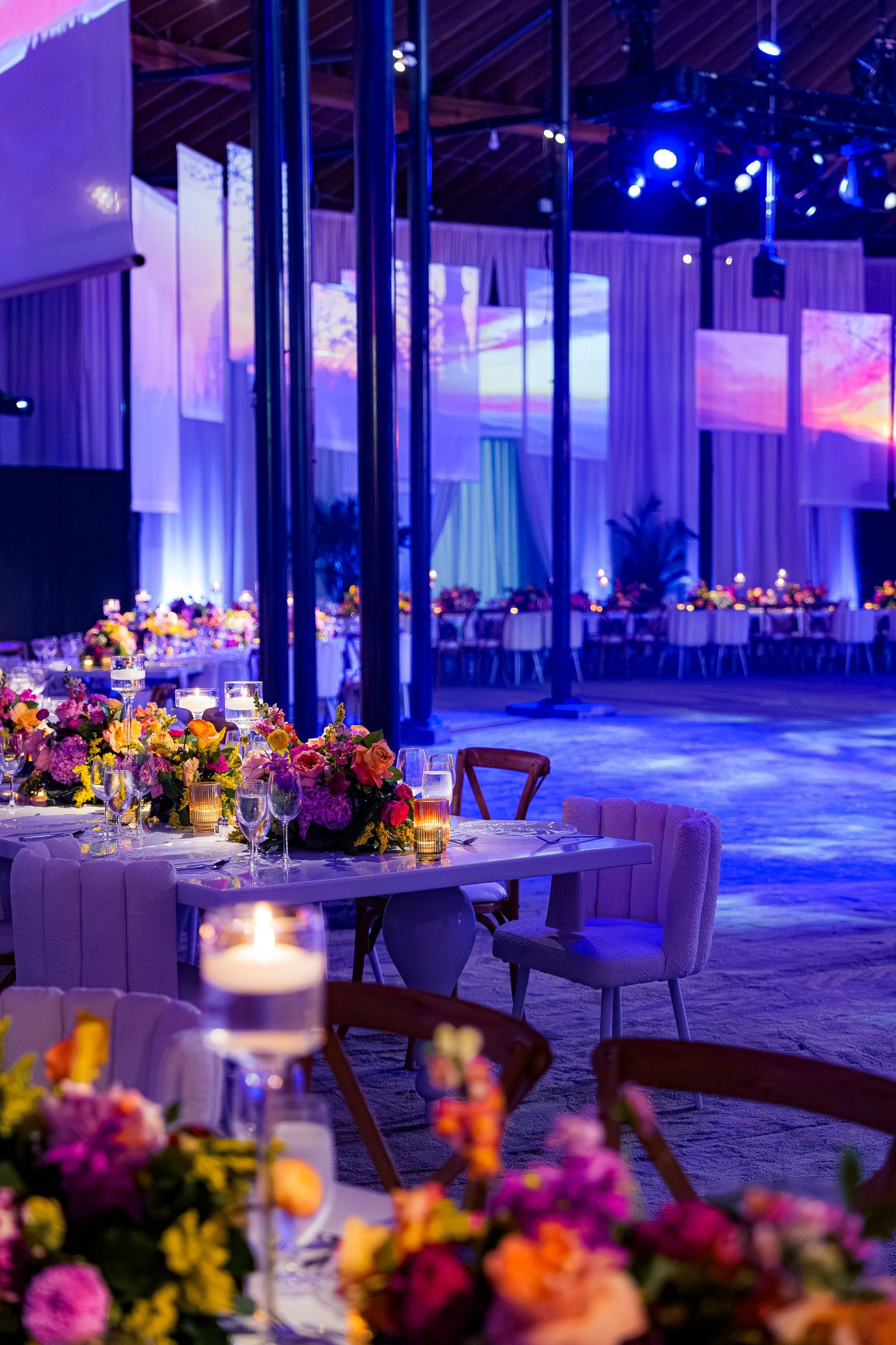 Modern event space with purple uplighting and elegant floral centerpieces on round dining tables.