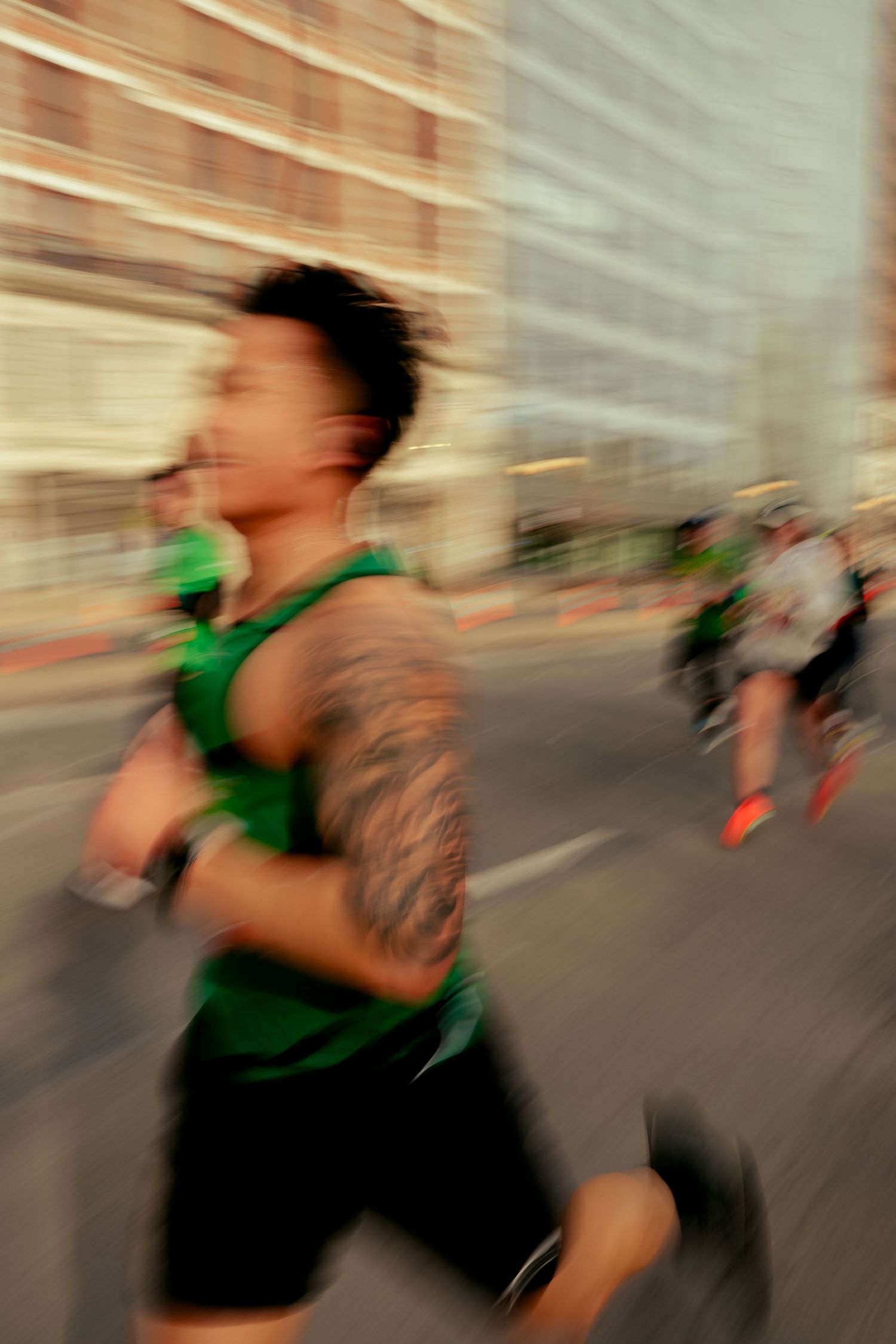A tattooed runner in a green tank top races through city streets during a marathon, captured with motion blur.
