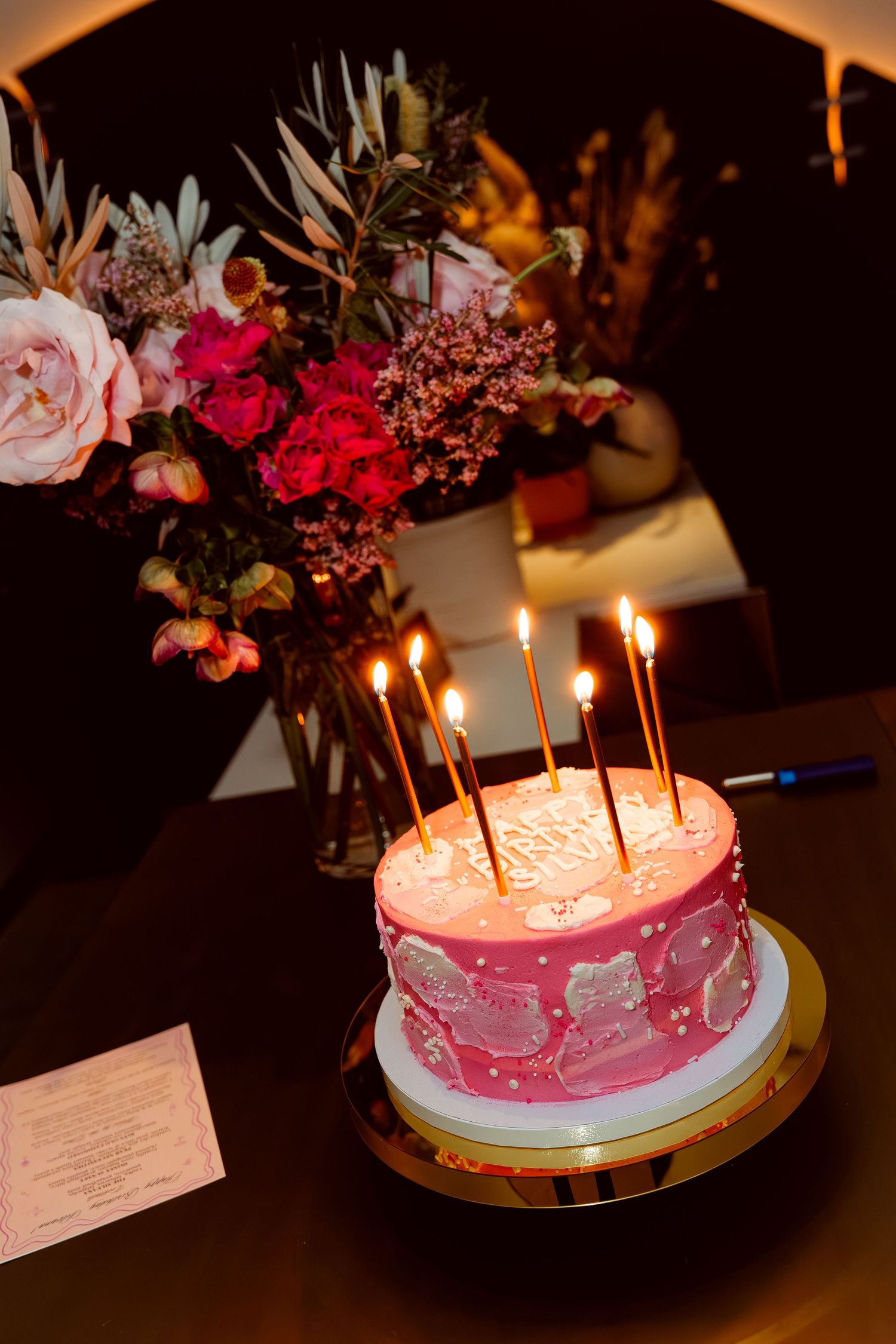 Pink birthday cake with lit candles and floral decorations on display.