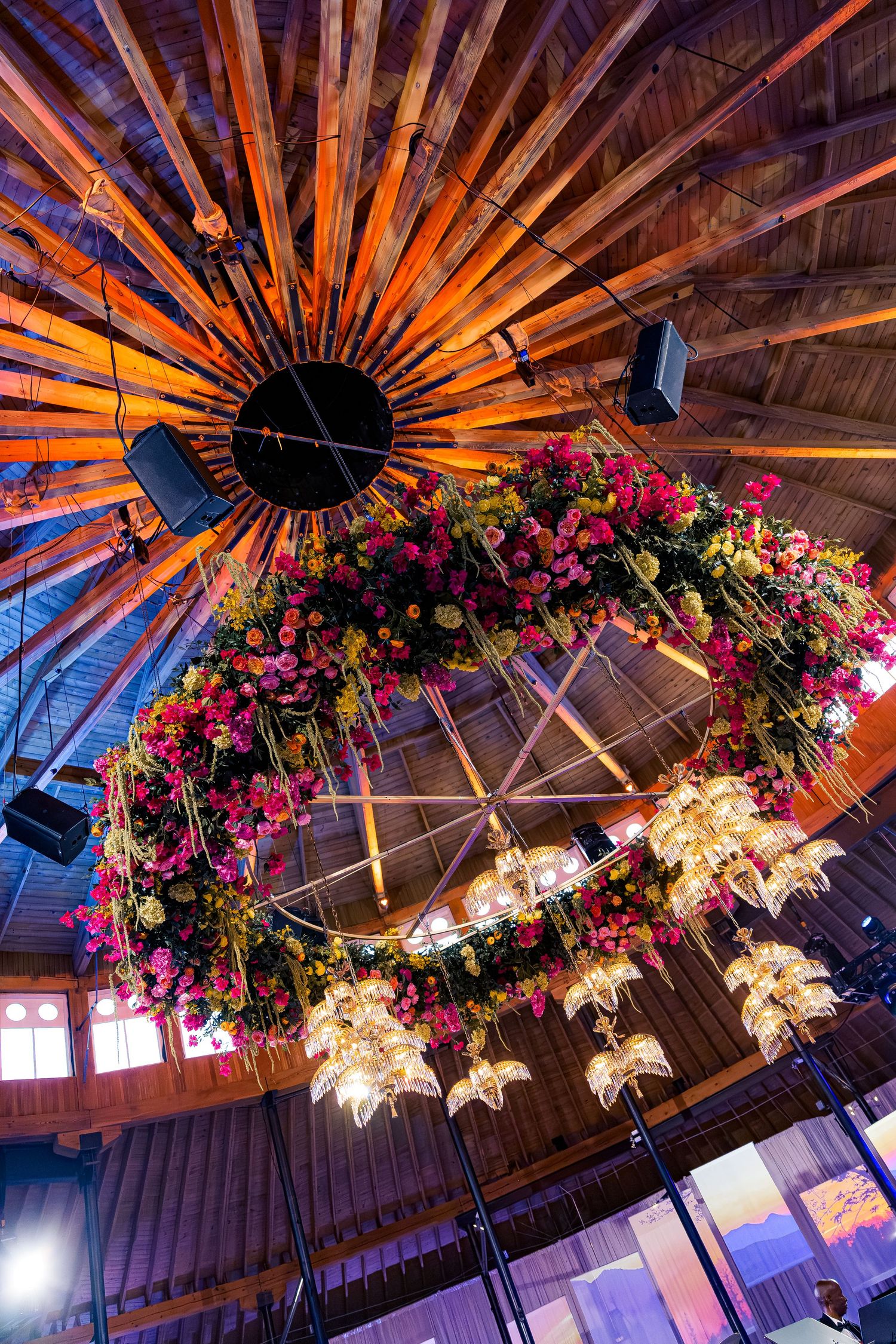 Ornate floral chandelier with pink flowers and wooden sunburst ceiling detail illuminated in warm lighting.