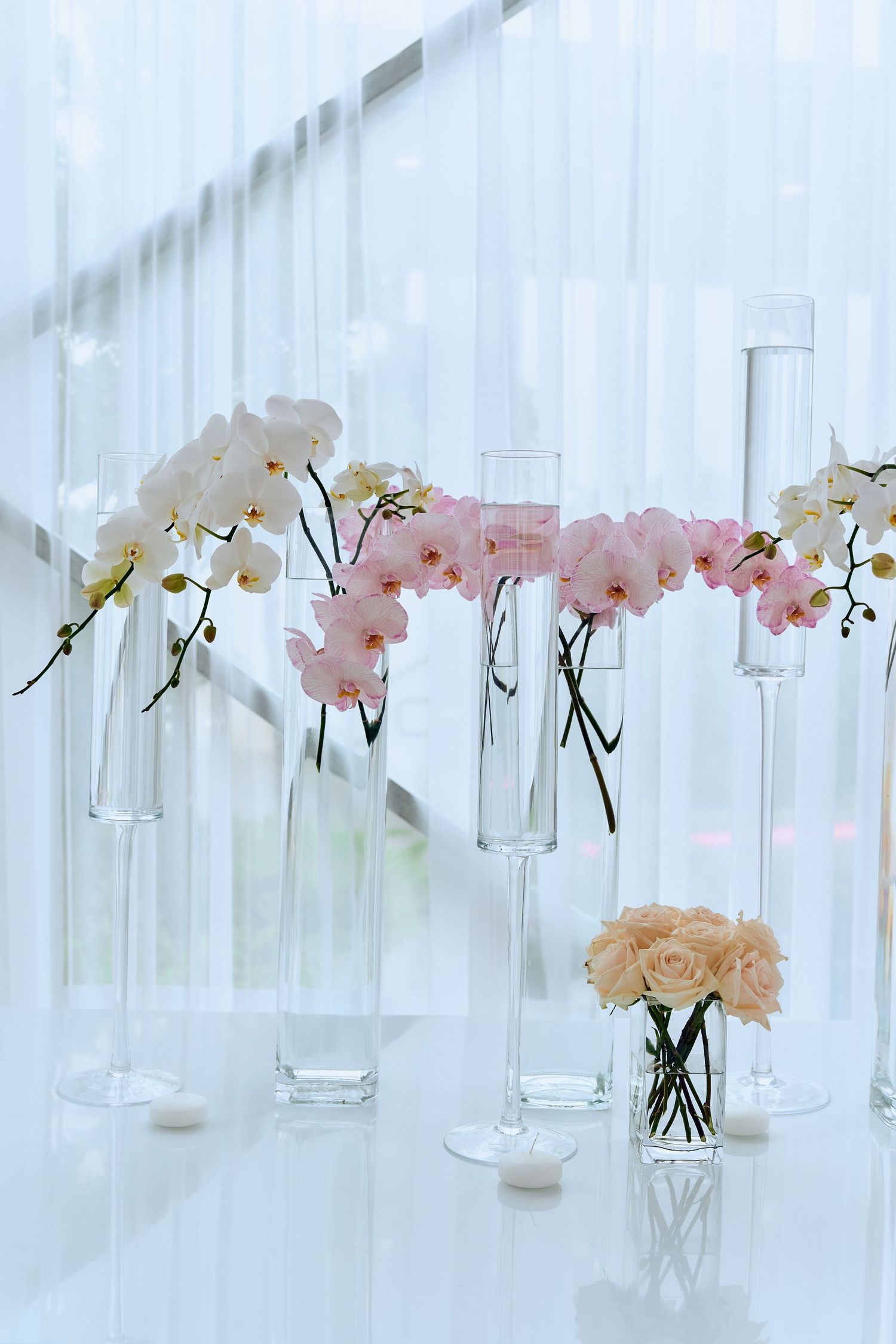 Minimalist white event decor featuring orchids and roses in clear glass vases.