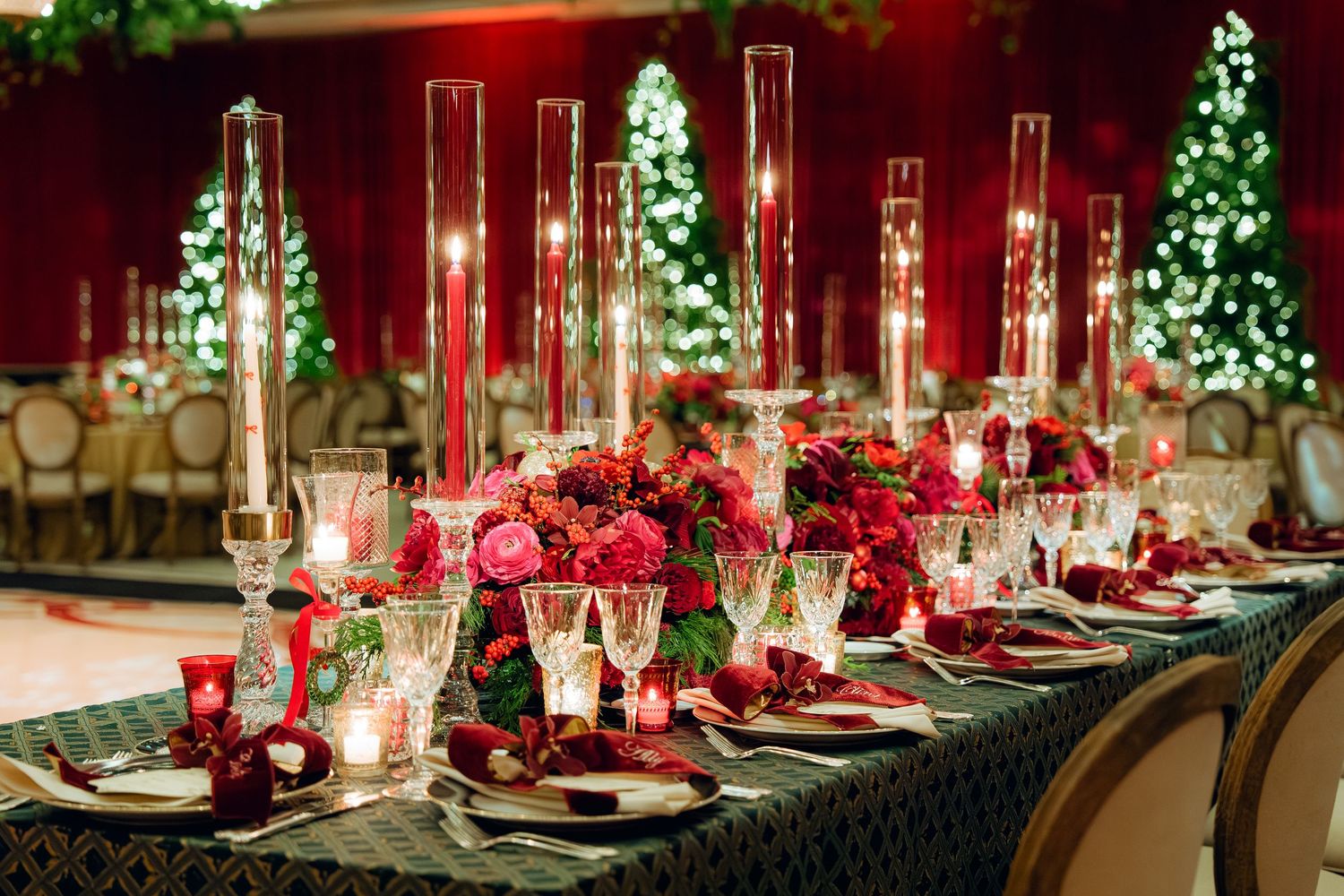 Formal table setting with red roses, crystal candleholders, and tall glass vases against red backdrop.
