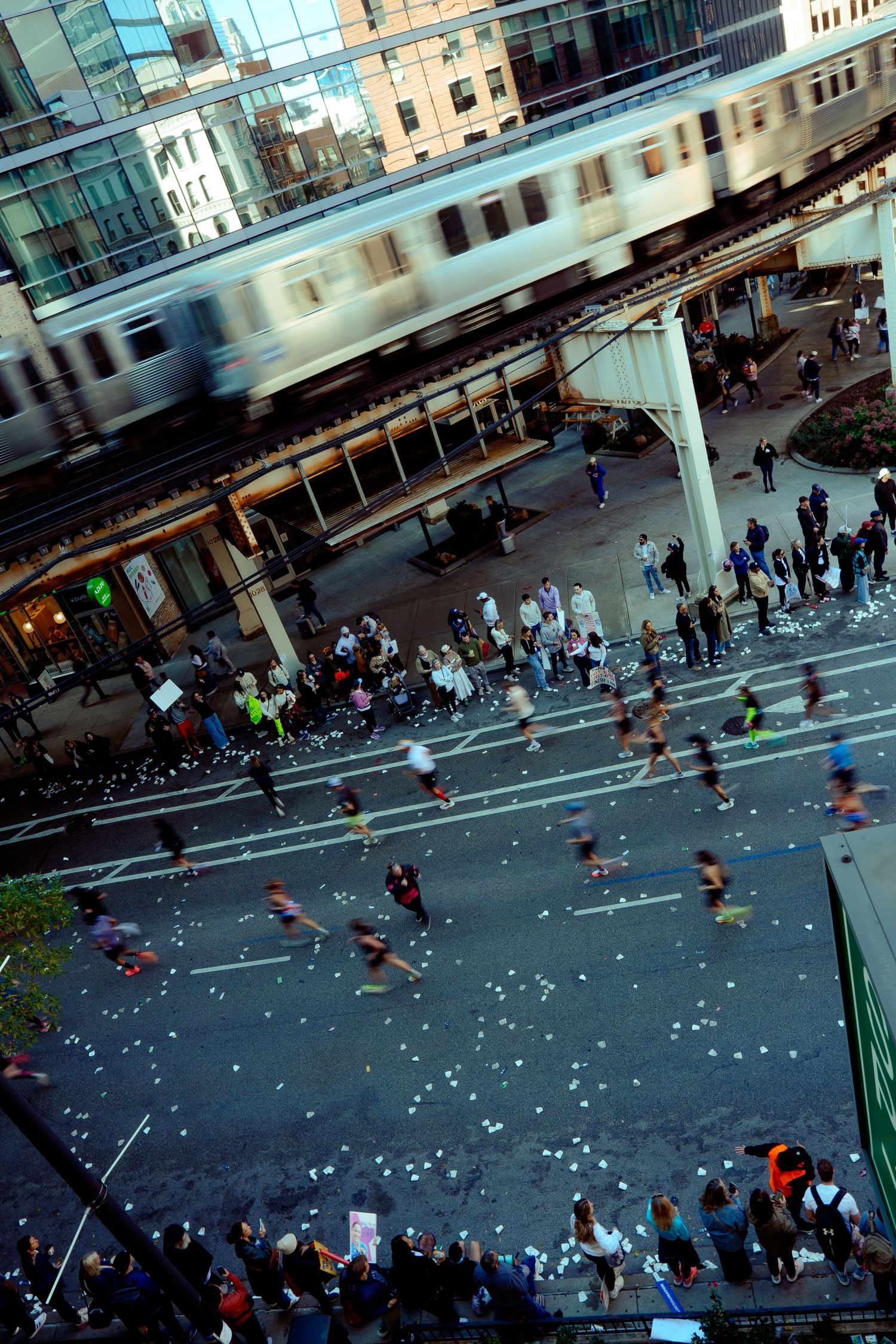 Elevated view of marathon runners passing under an elevated train track in a city.