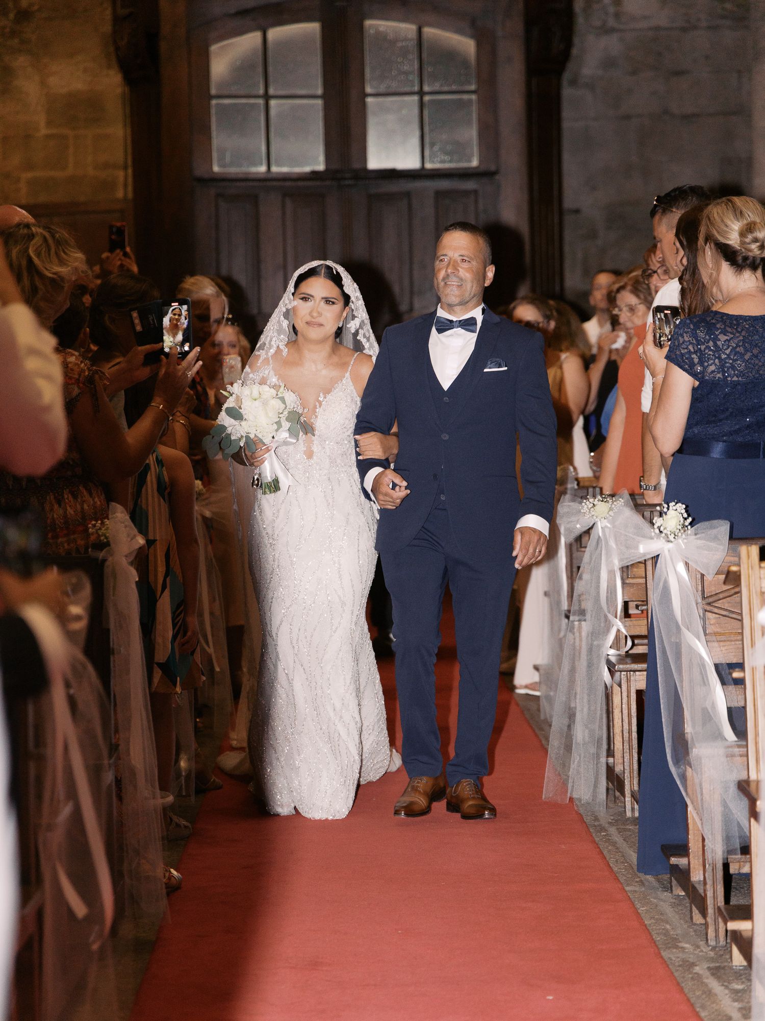 A wedding couple walks down a church aisle lined with guests while photographers capture the moment.