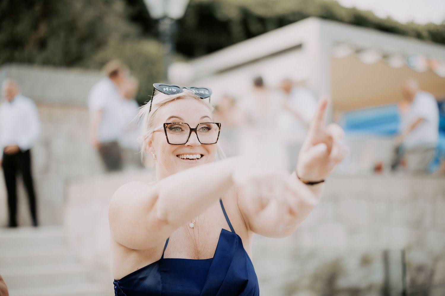 Person in a navy blue dress and glasses extends their arms toward the camera in a playful outdoor pose.