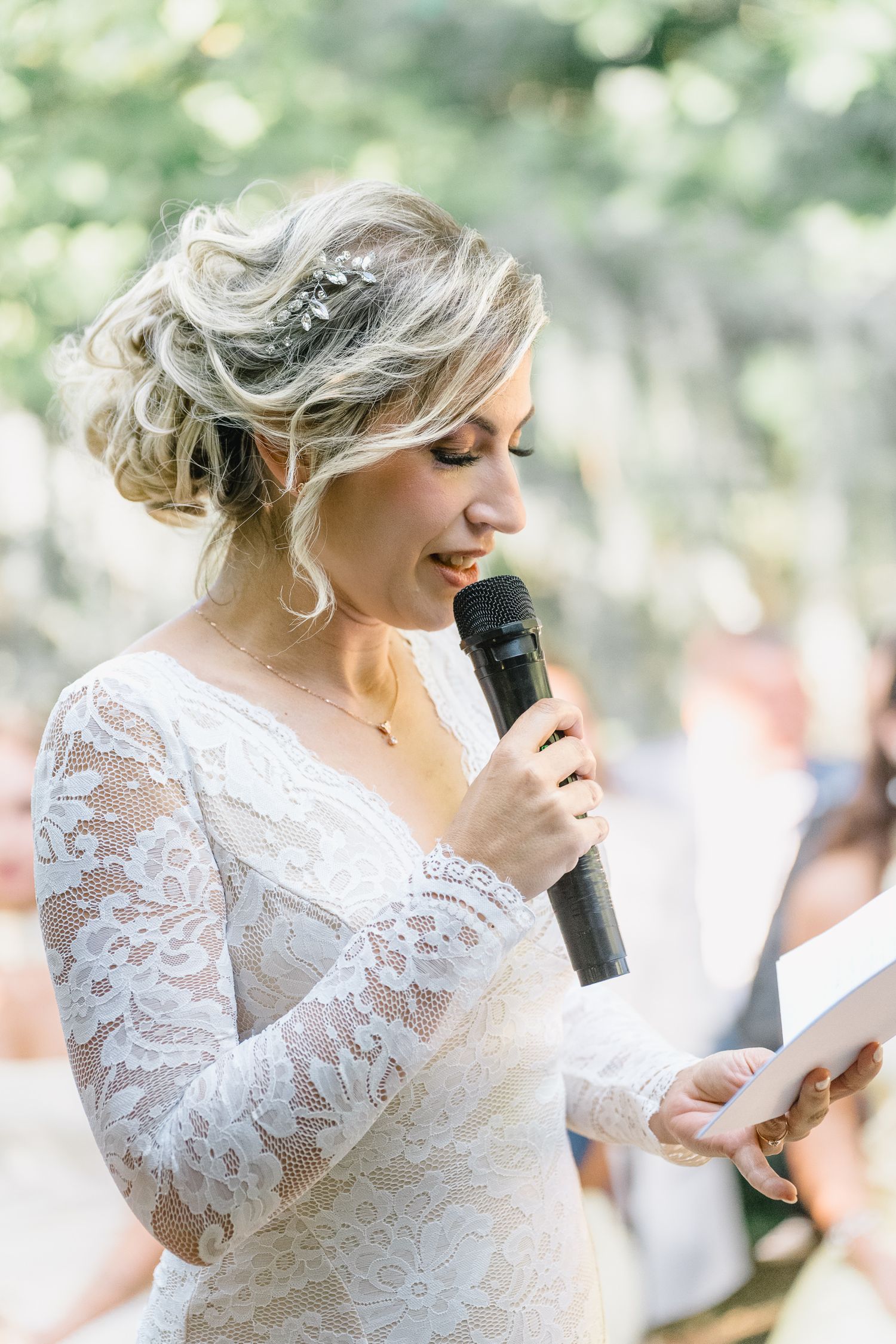 A bride in a long-sleeve lace wedding dress reads from paper while speaking into a microphone during an outdoor ceremony.