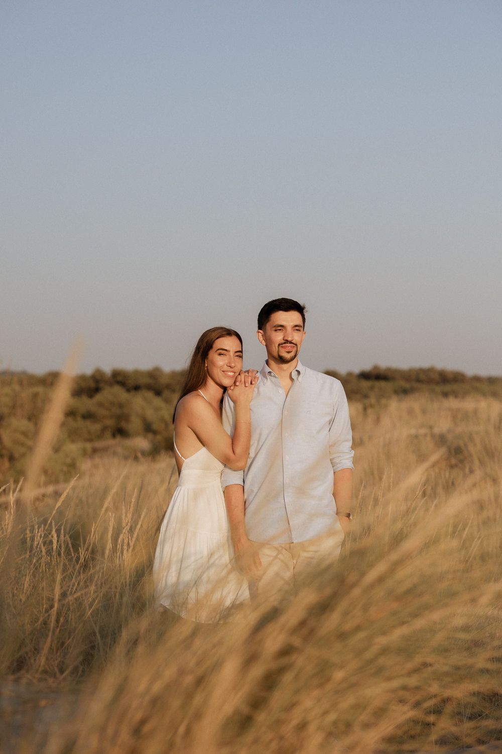 Um casal romântico posa junto em um campo dourado ao pôr do sol, vestindo roupas brancas.