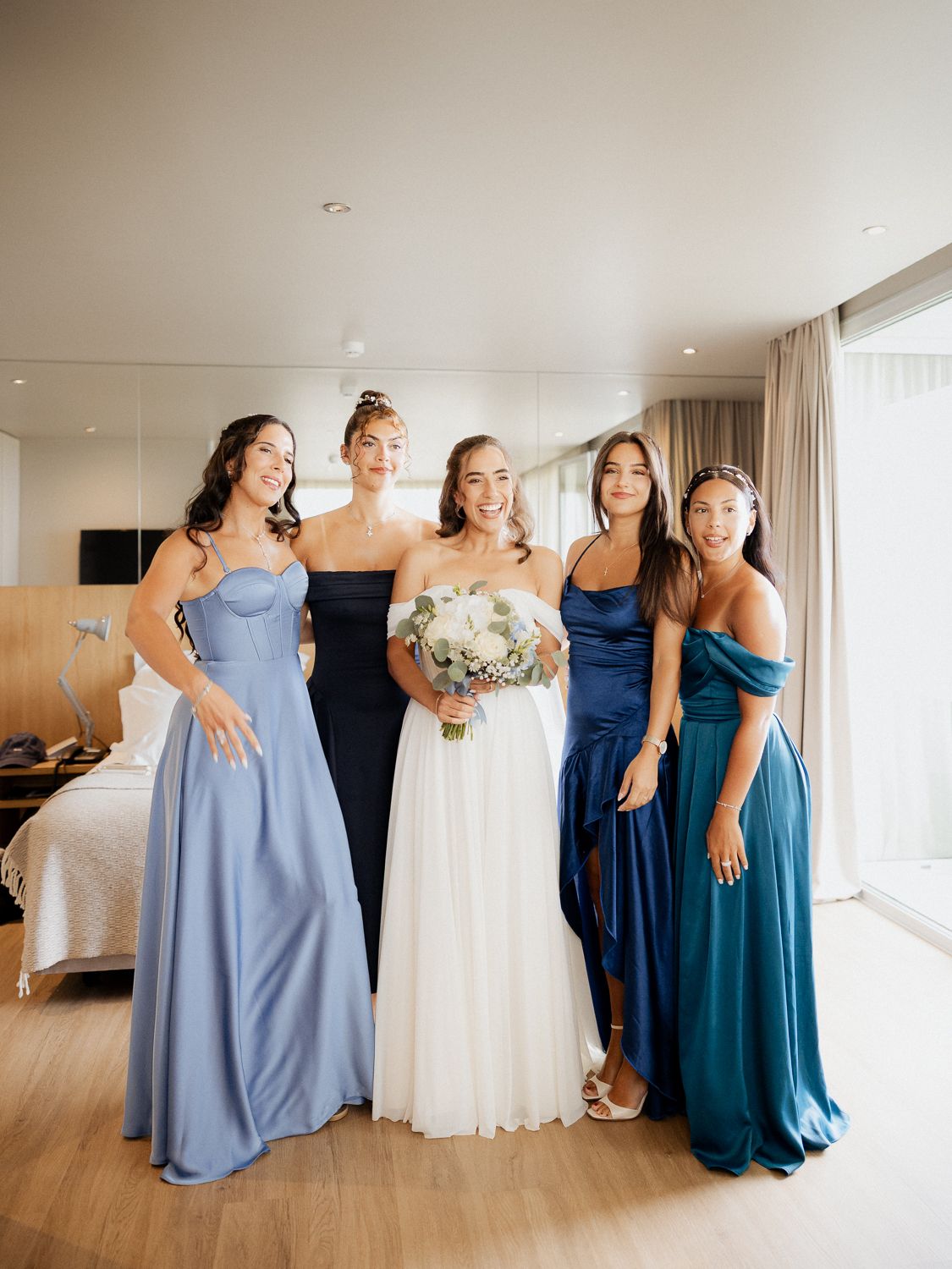 Wedding party in coordinating blue dresses and white gown pose together in bright indoor venue.