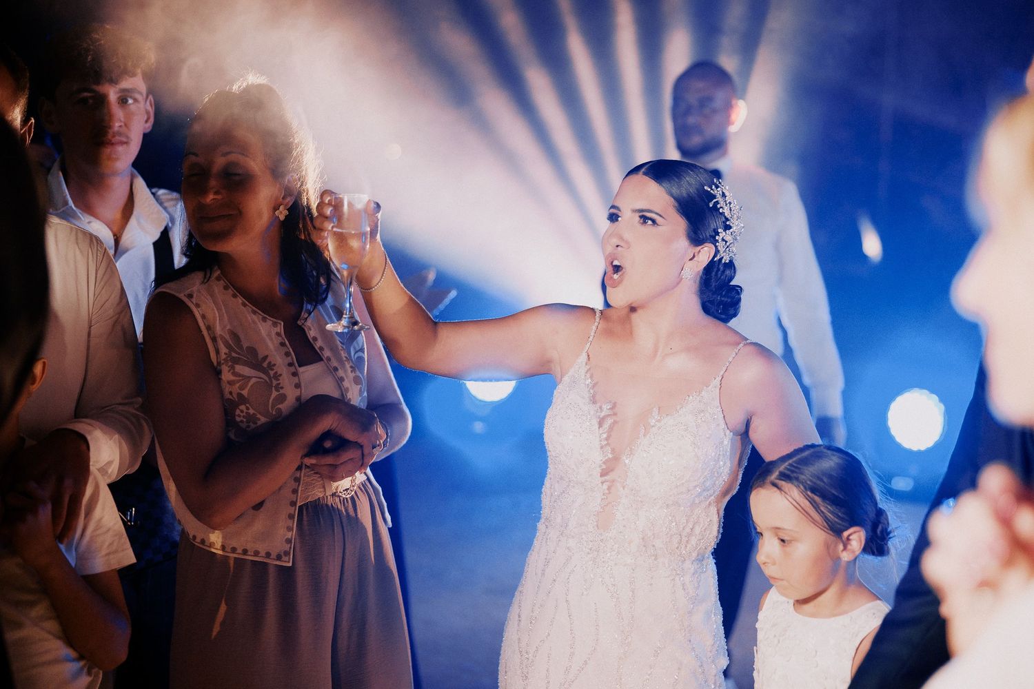 Wedding guests dance and celebrate together on a dimly lit dance floor with blue lighting effects in the background.