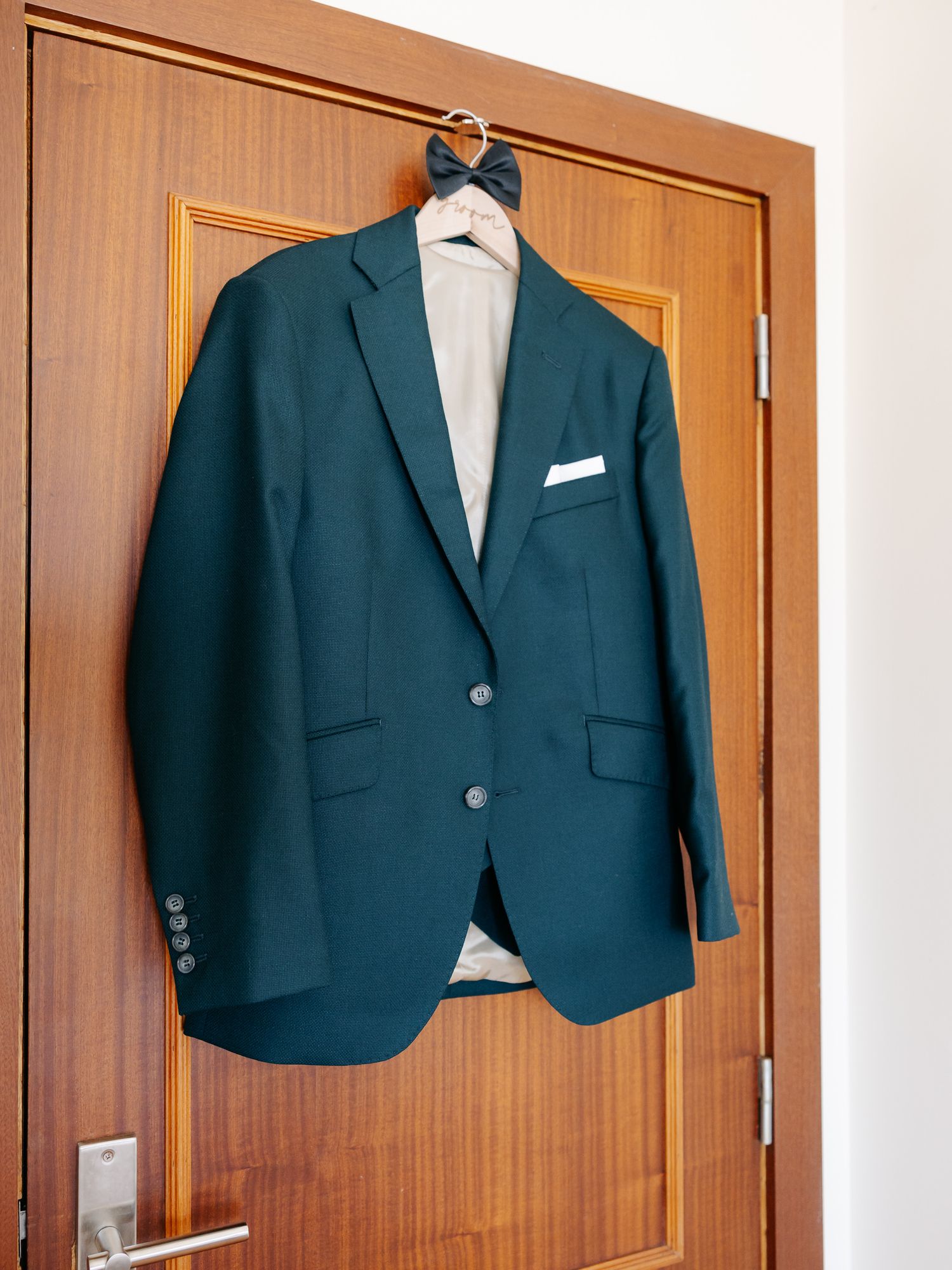 A navy blue suit jacket hangs on a wooden door against a light colored wall.