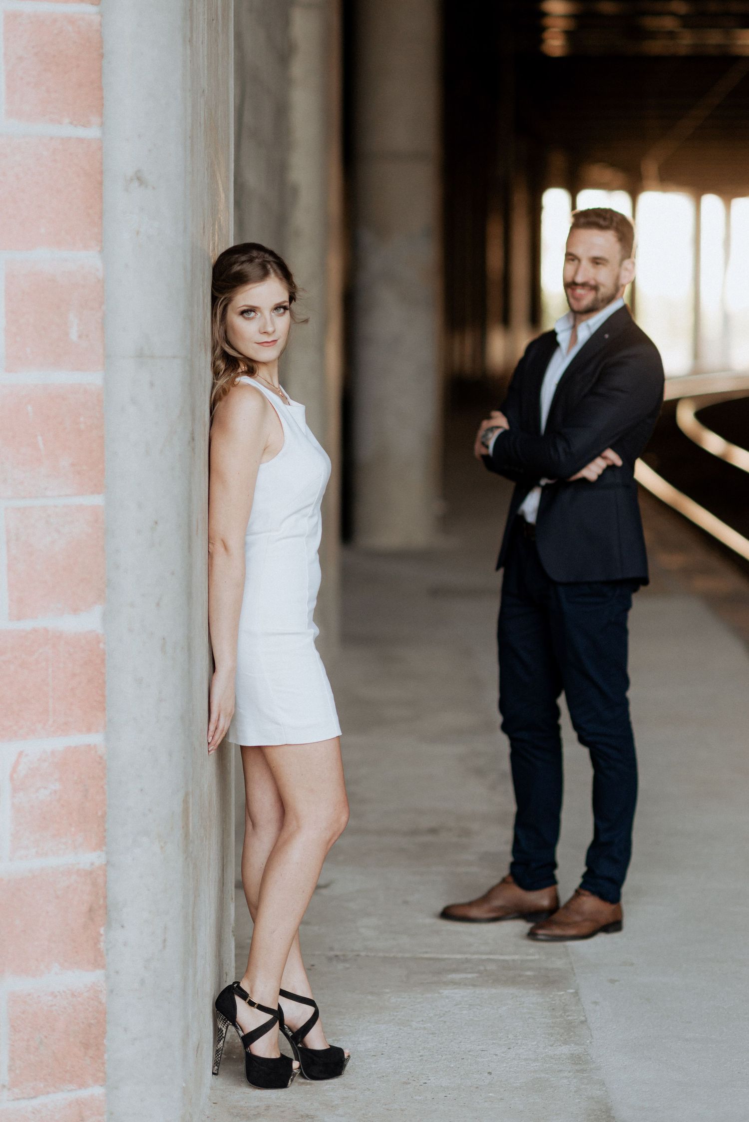 Um casal posa em um cenário urbano moderno, a mulher com um vestido branco e o homem com um terno escuro.