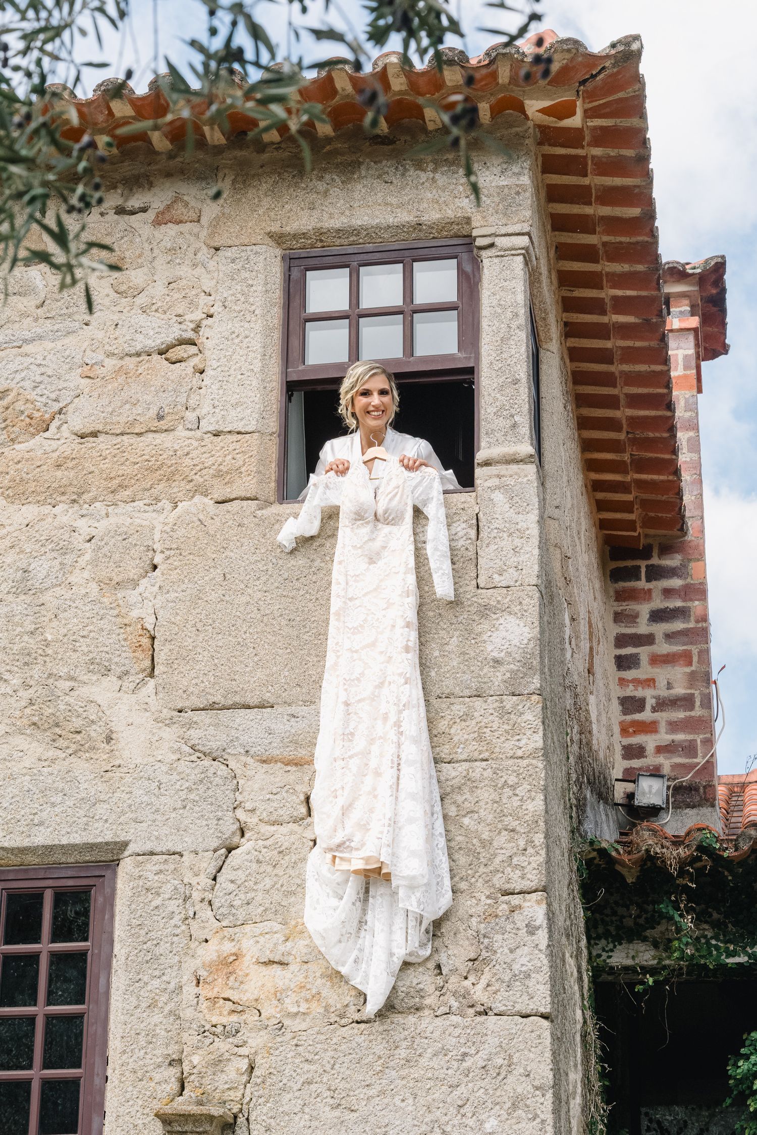 A white vintage dress hangs on an old stone wall with brick details and a window frame in a rustic setting.