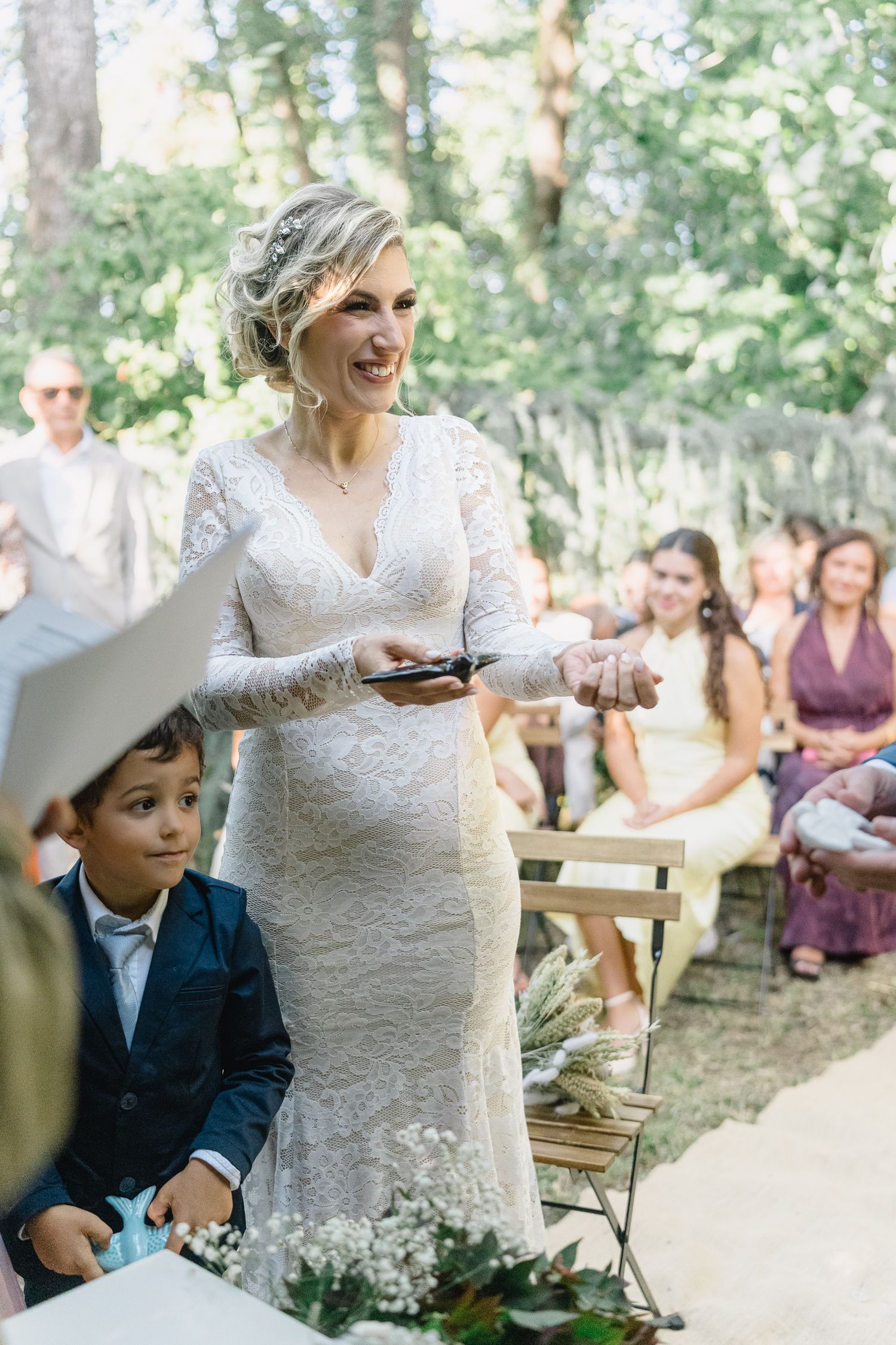 Outdoor wedding scene showing guests seated in a wooded area as a bride in lace cuts a rustic cake.