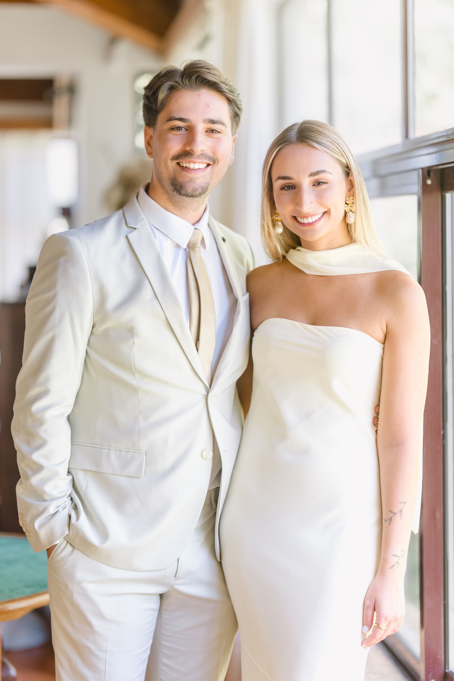 A couple in matching white formal attire poses together at a wedding venue with natural lighting.