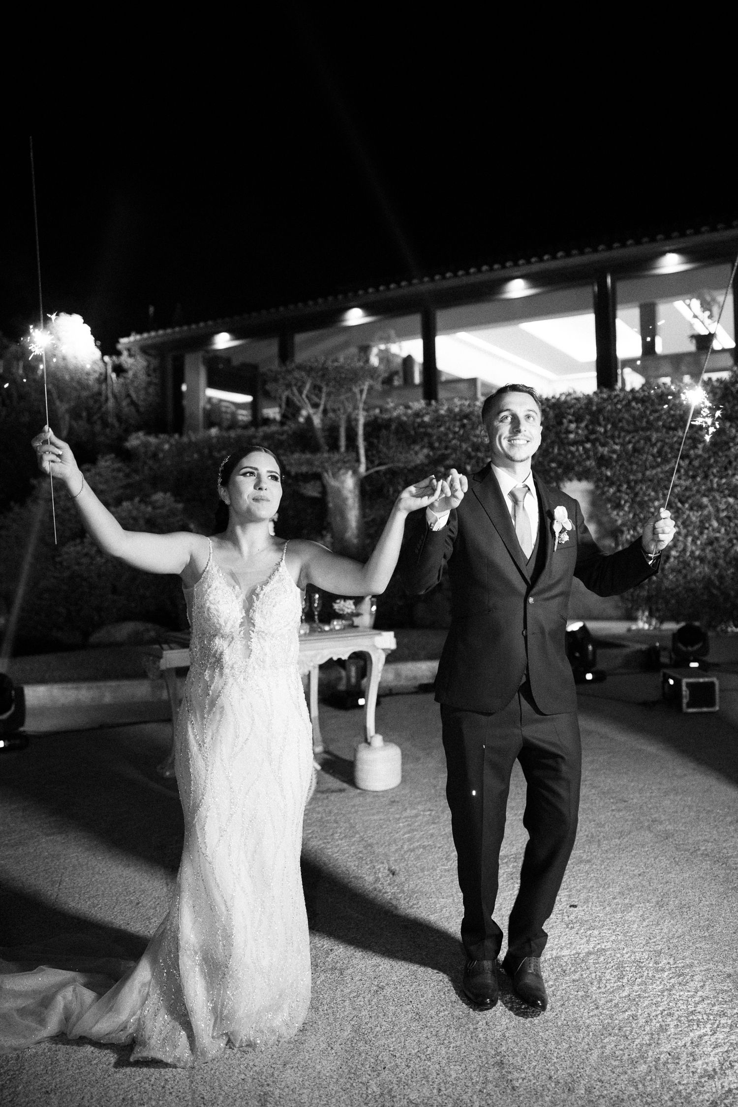 A couple celebrates their wedding night holding sparklers while dancing outdoors at an illuminated venue.