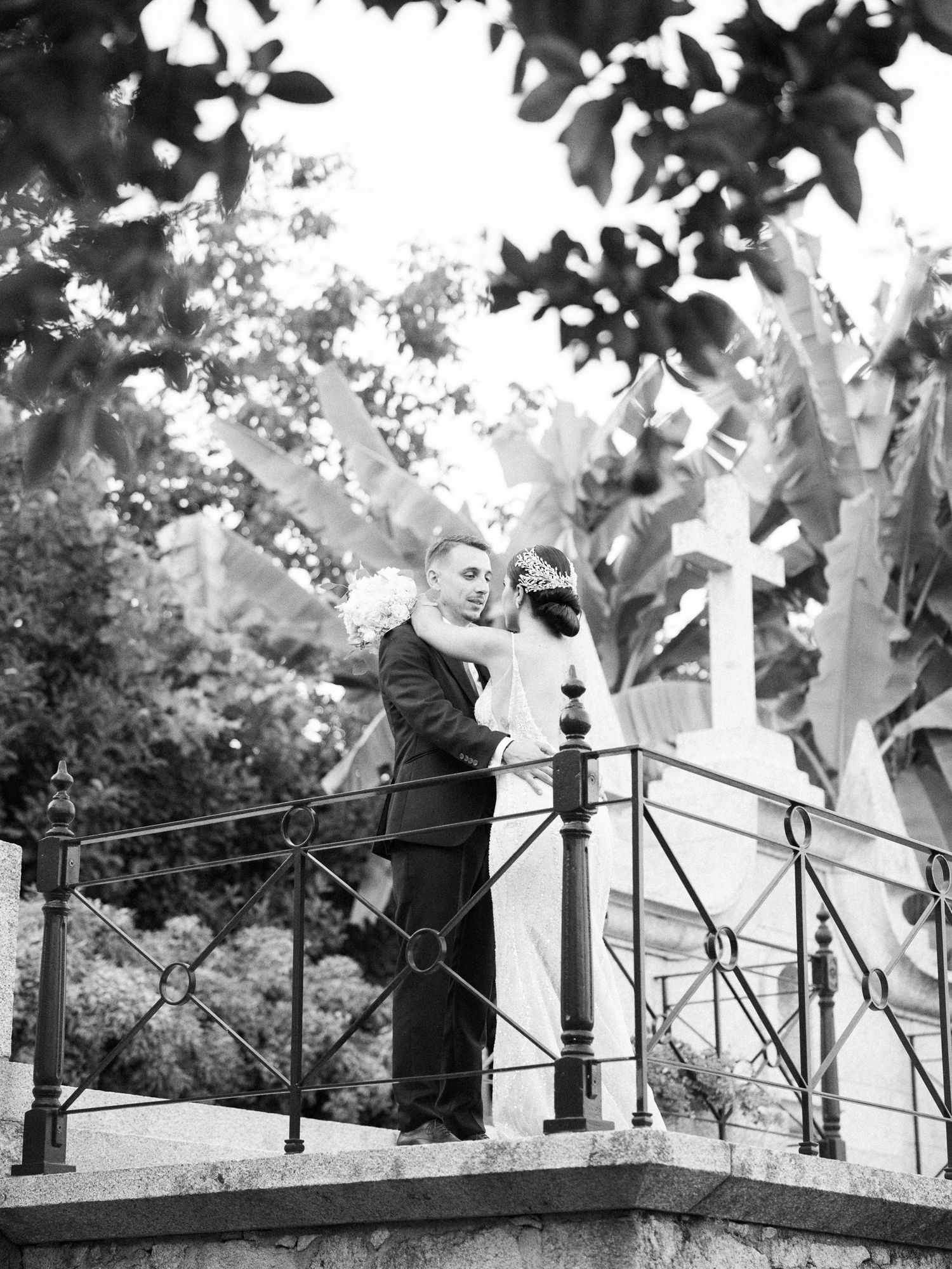 A black and white artistic wedding photograph showing a couple on a stone balcony with trees in the background.