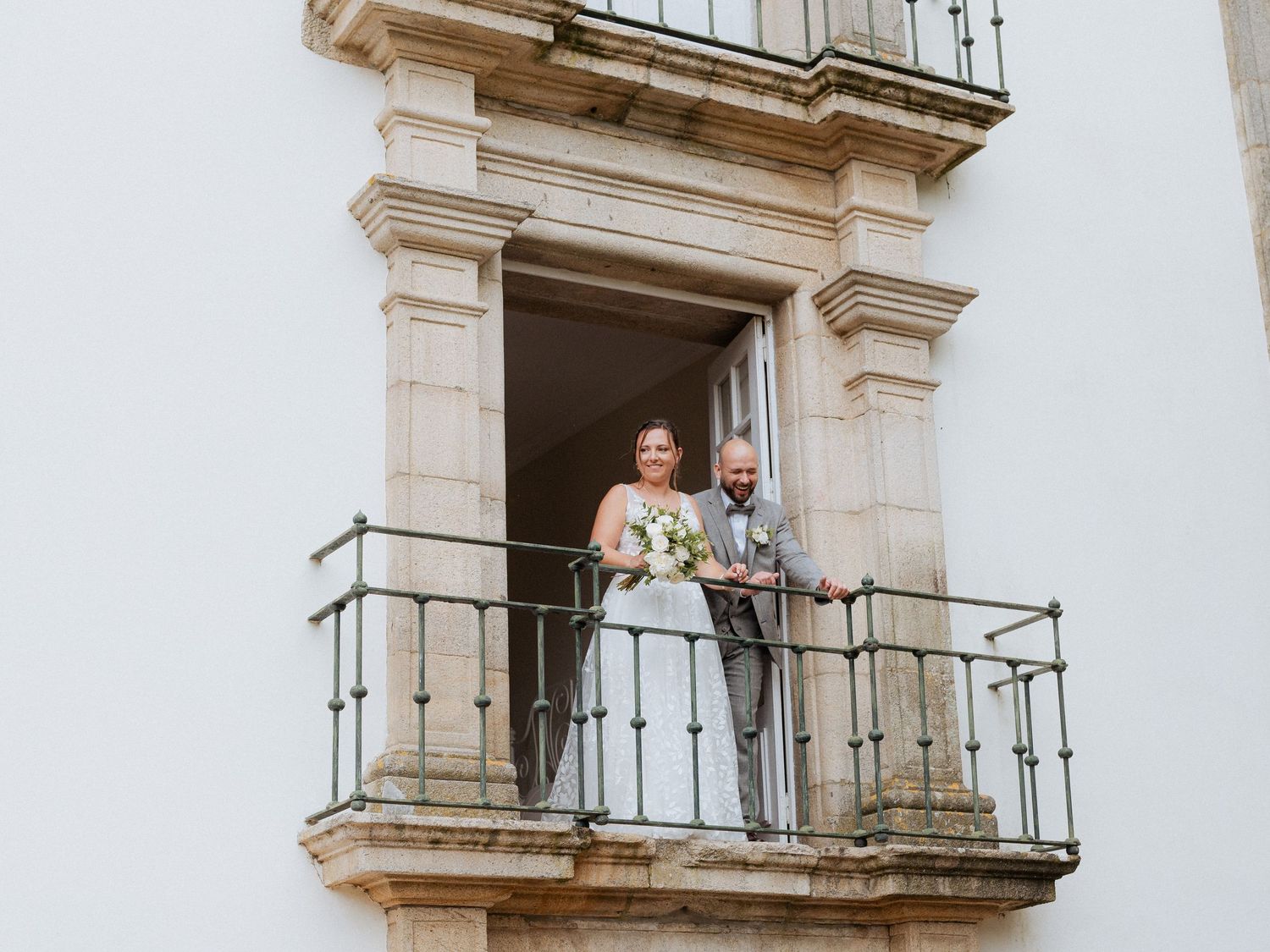 A couple stands together on an ornate stone balcony of a white building during their wedding celebration.