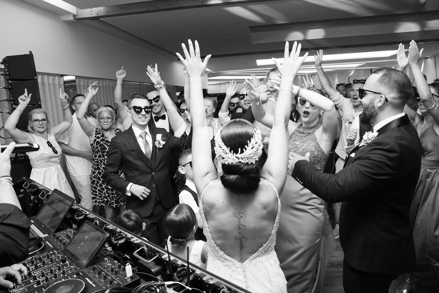 Black and white photo of a lively dance party with raised hands and a DJ setup in a crowded room.
