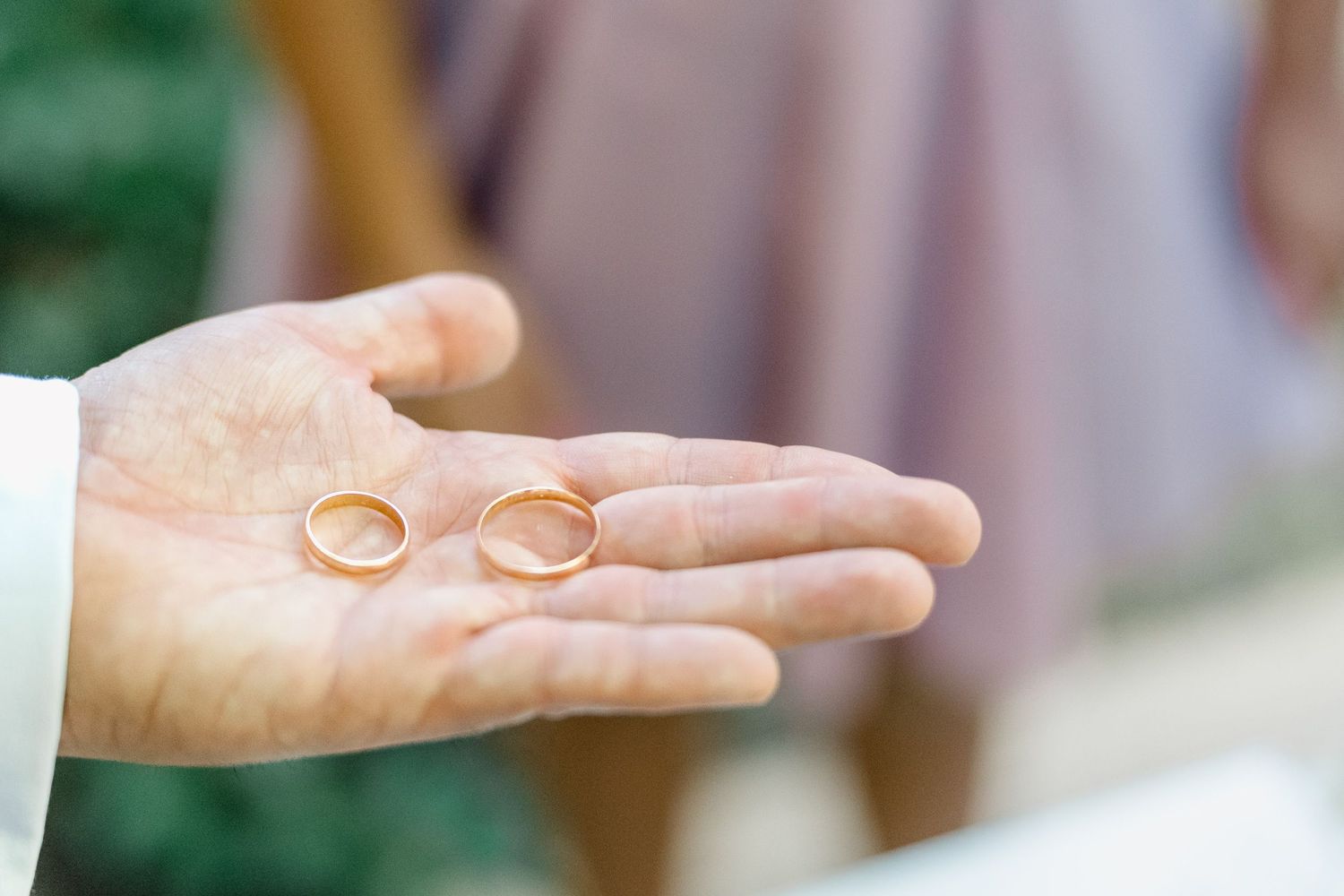 Uma mão segura duas alianças de casamento em ponte de lima, douradas contra um fundo suave e desfocado em tons de verde e rosa.