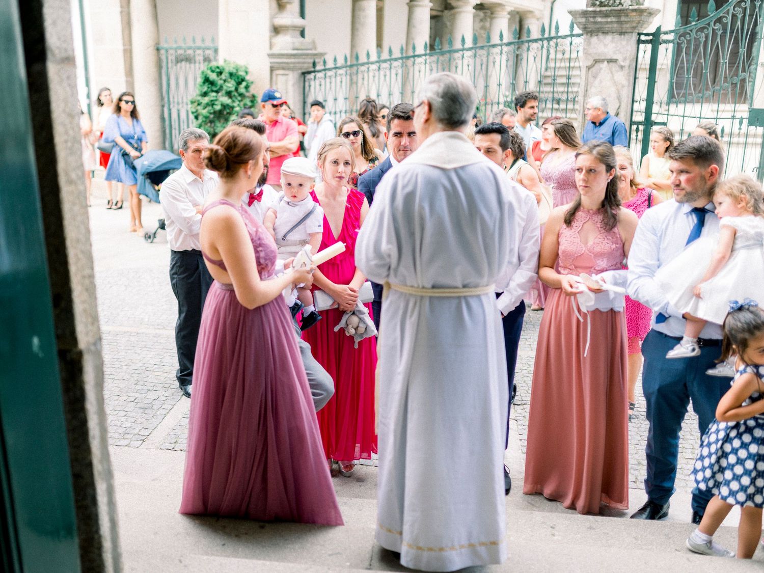 A group of wedding guests in formal attire gather around a clergy member in white robes outside a church entrance.