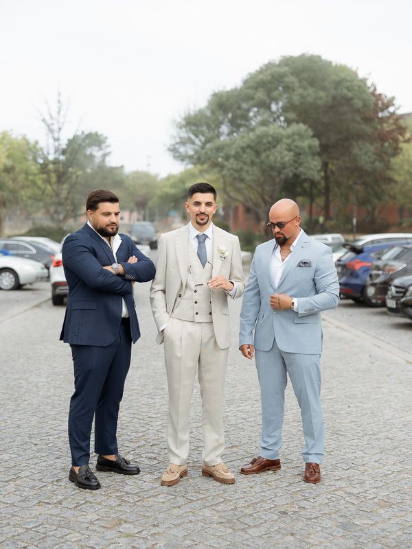 Three well-dressed men in suits posing in a cobblestone parking lot, wearing navy, beige, and light blue formal attire.
