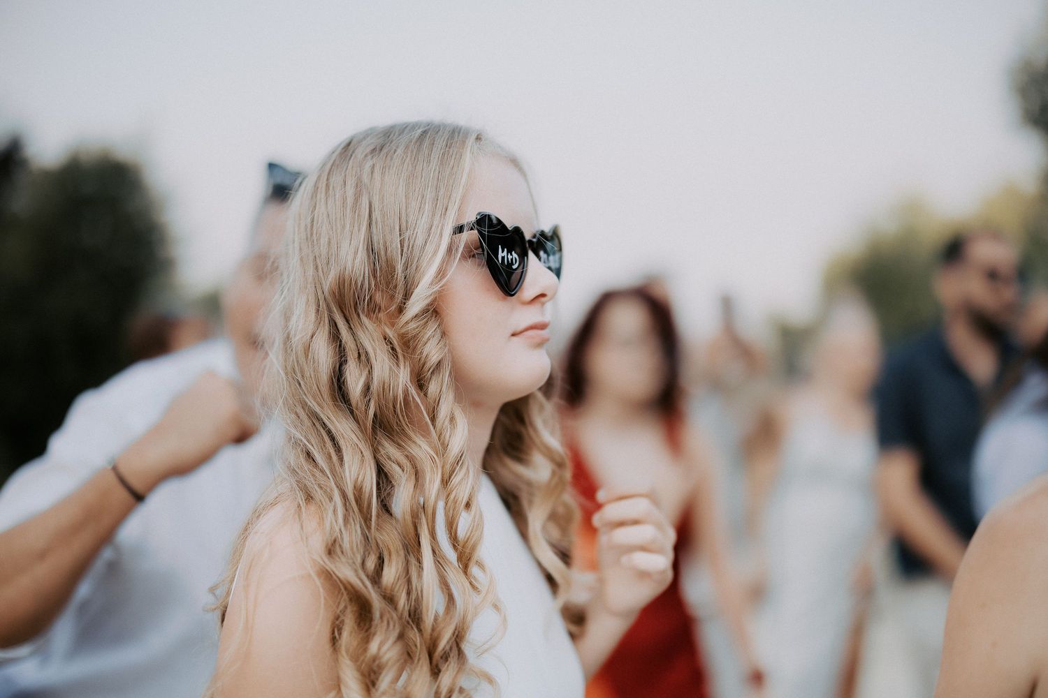 A person with long blonde wavy hair and sunglasses smiles at an outdoor gathering with others in the background.