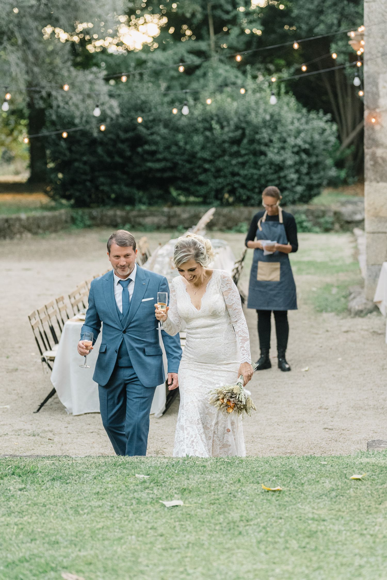 A couple walks together at an outdoor wedding ceremony with string lights and greenery in the background.