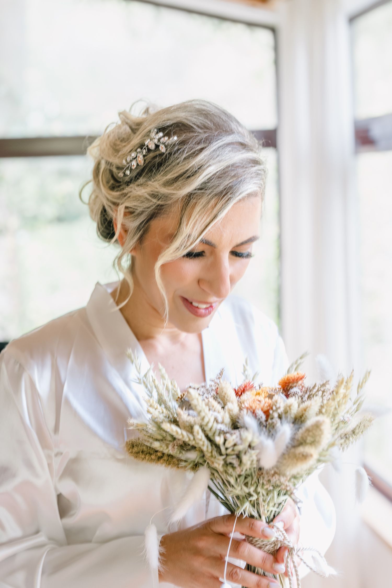 Bride in white robe smiles while holding dried floral bouquet in sunlit room during wedding preparations.