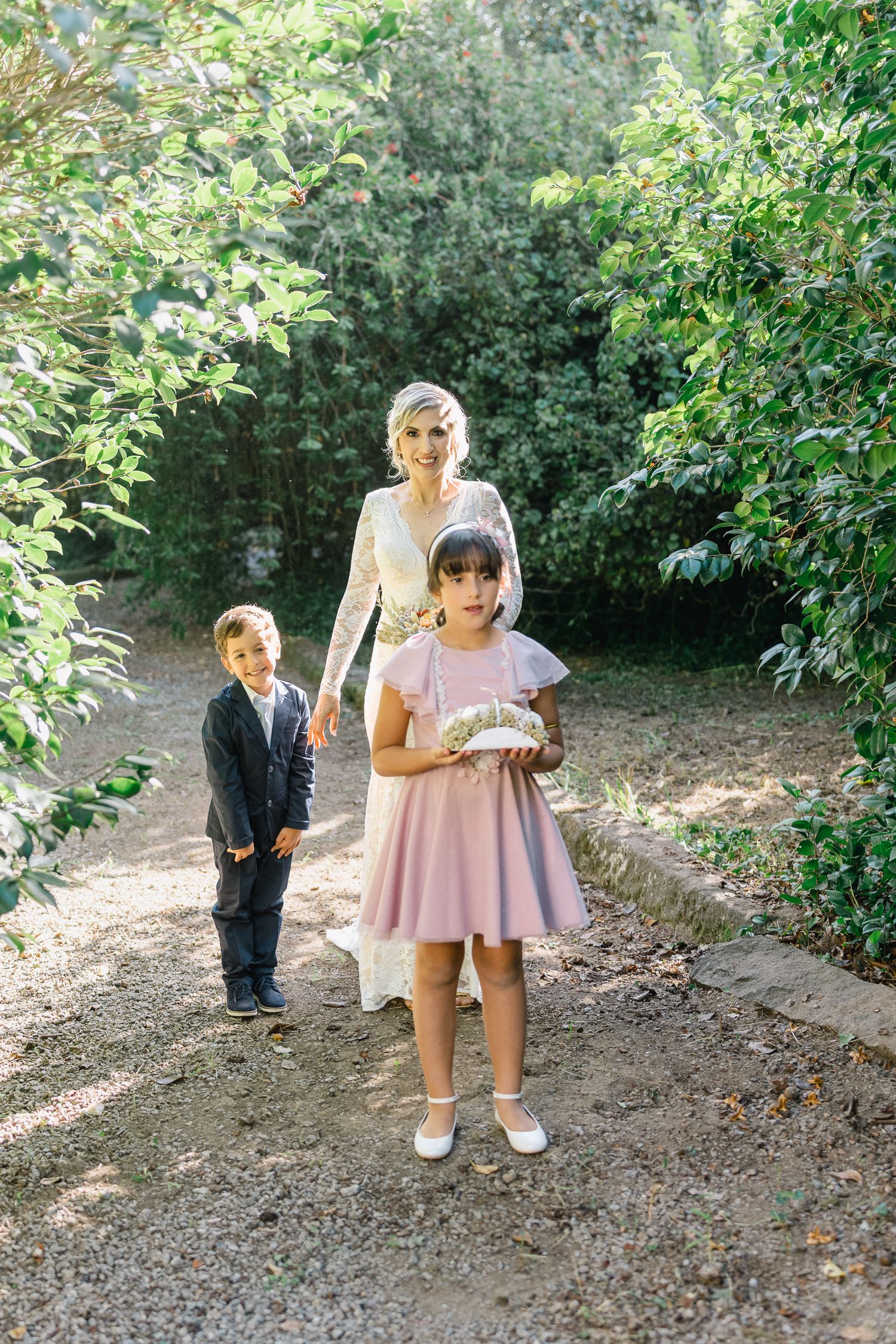 Small children in formal attire playing and exploring in a garden path surrounded by green foliage.