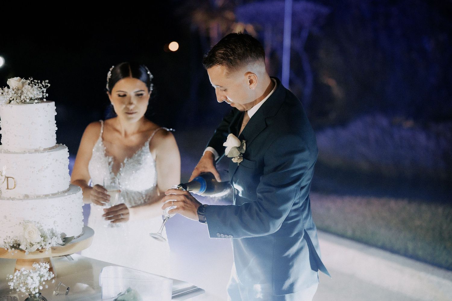 A couple cuts their elegant white wedding cake together during an evening reception celebration.