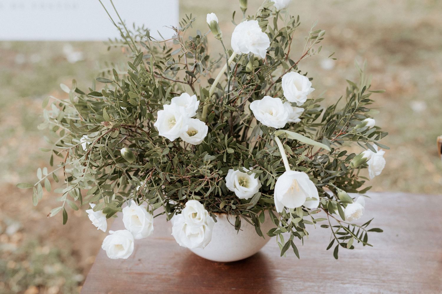 Rosas brancas e folhagens delicadas dispostas em um vaso de cerâmica cor creme sobre uma superfície de madeira. Casamento na Quinta da Pia