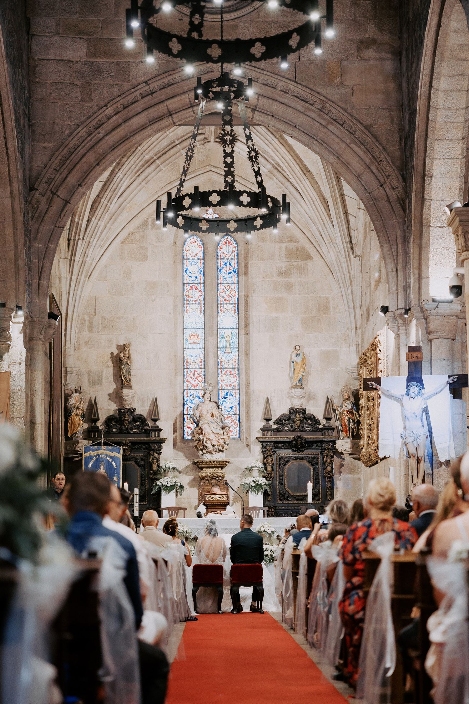 Interior view of a church wedding ceremony with stone arches, stained glass windows and ornate chandeliers hanging above.