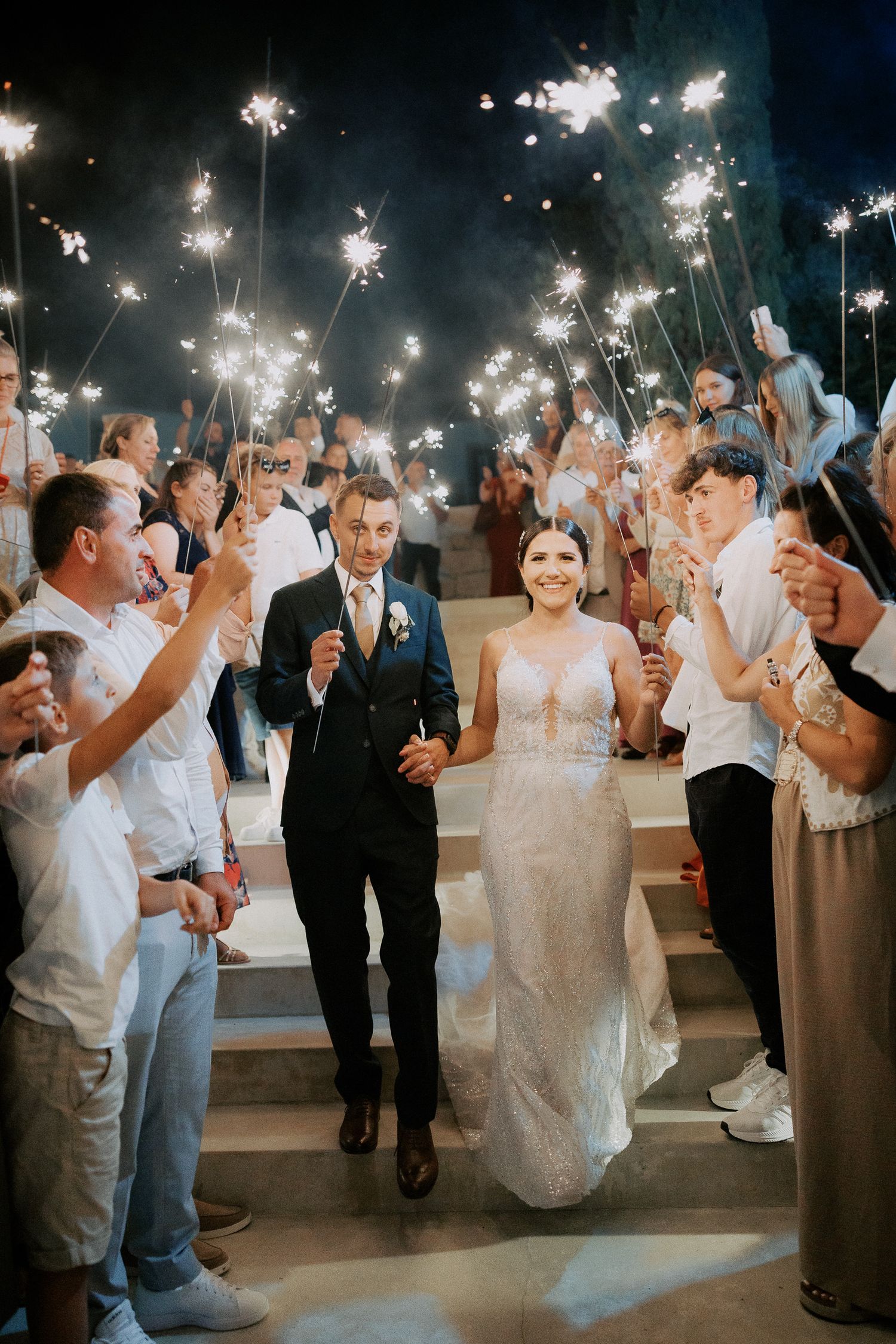 Wedding guests hold sparklers during an evening celebration as newlyweds dance in a romantic outdoor reception.