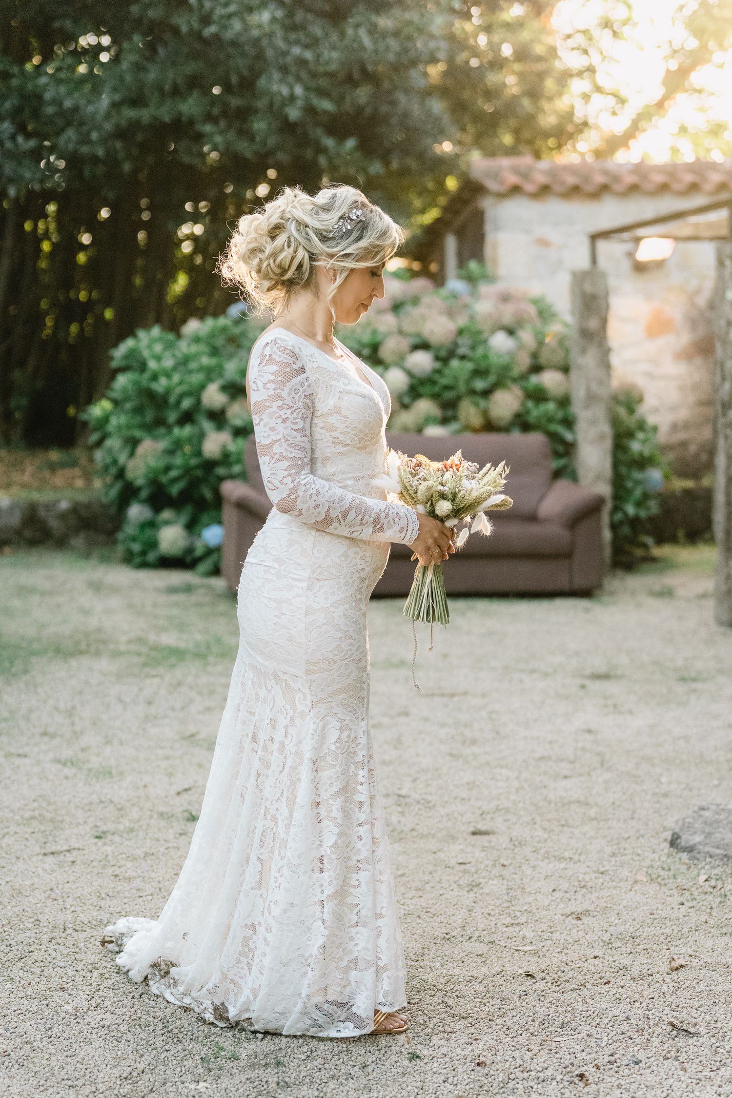 Uma noiva com um vestido de renda de mangas compridas segura um bouquet num jardim ao ar livre durante a hora dourada em ponte de lima