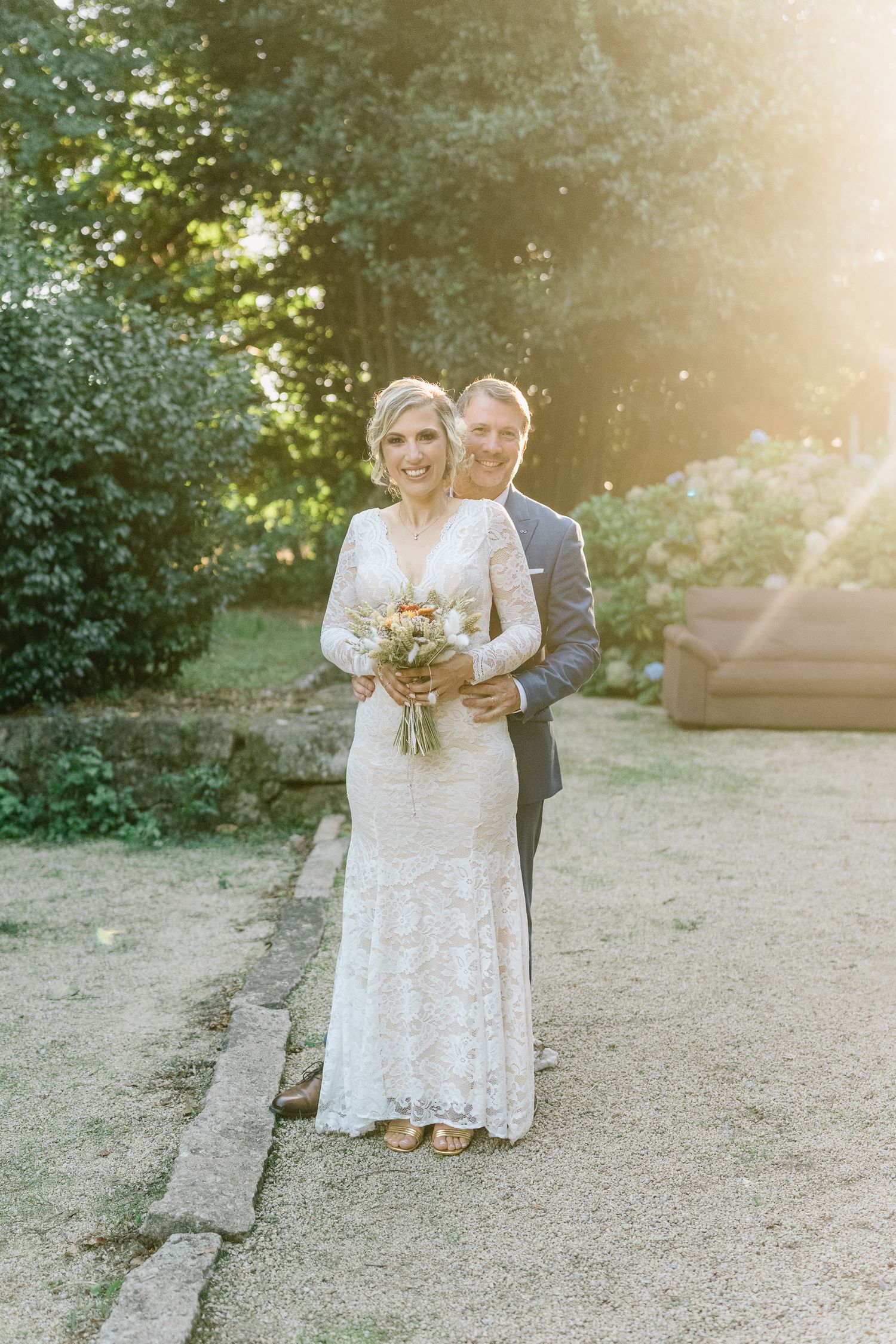 Um casal com trajes de casamento posa ao ar livre sob a luz dourada do sol, com uma paisagem natural de jardim como pano de fundo.