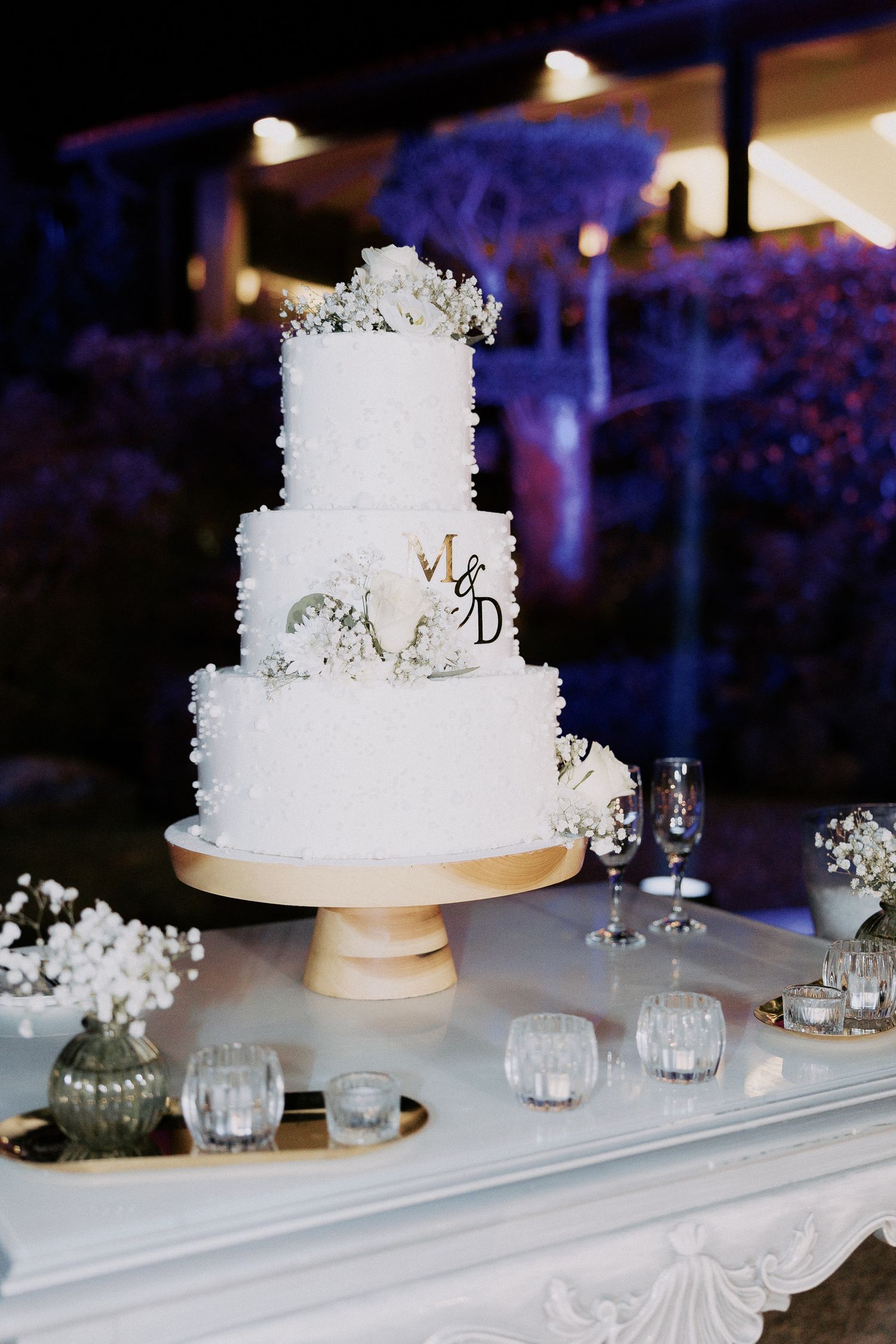 White wedding cake with delicate floral details sits on gold cake stand surrounded by glass votives and baby's breath.