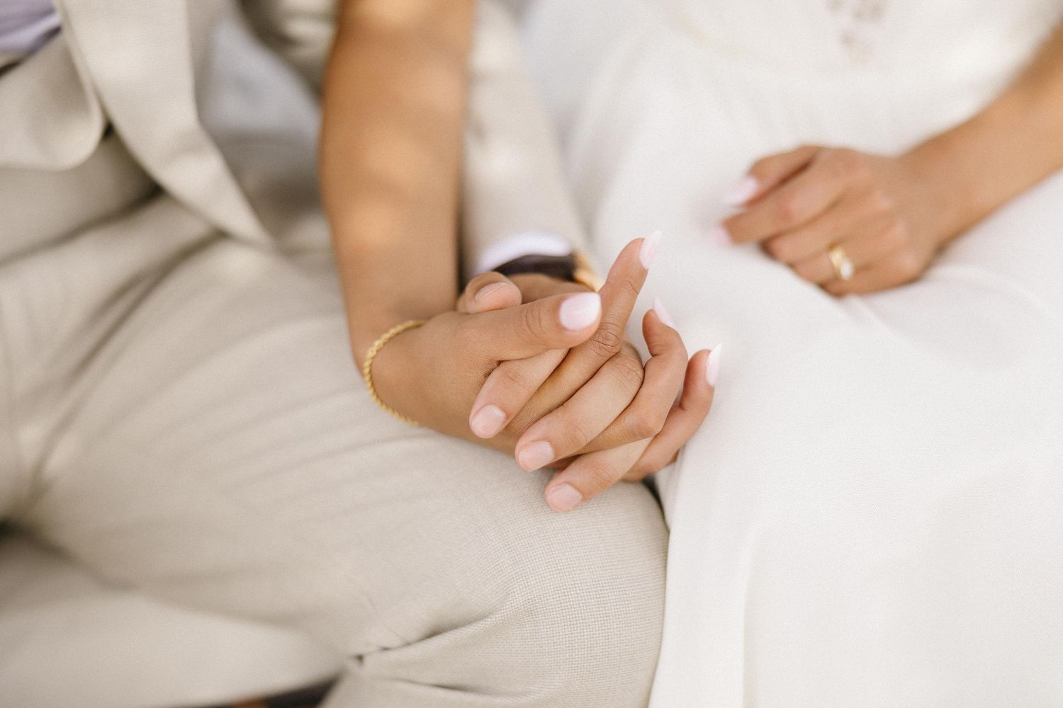 Close-up de mãos entrelaçadas mostrando alianças de casamento contra trajes formais brancos e bege.