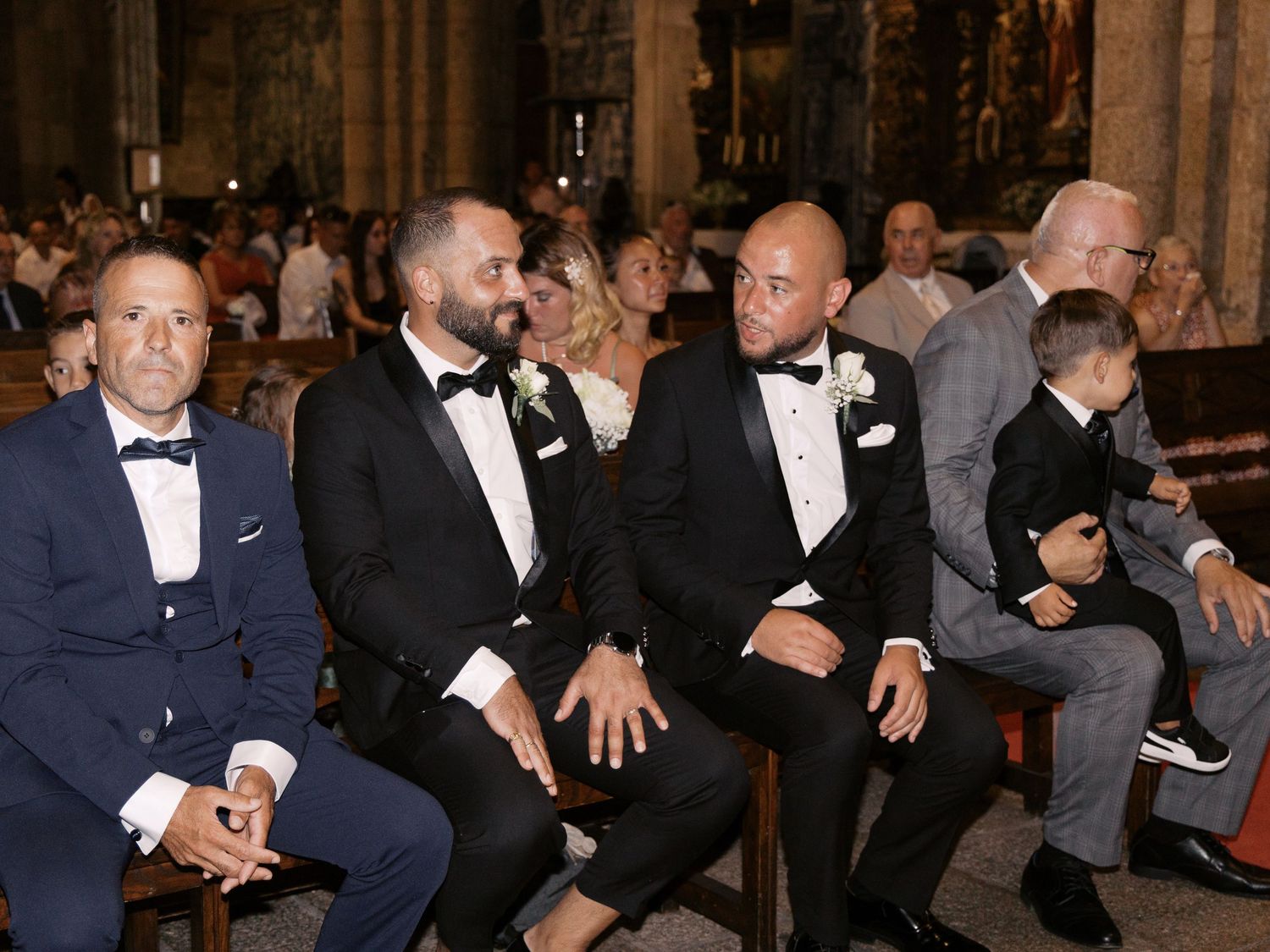 A row of formally dressed people in black tuxedos and suits seated in an elegant venue during an event.