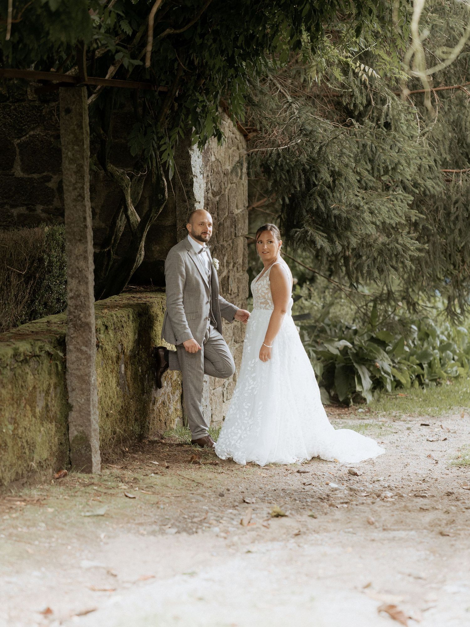 A romantic couple in wedding attire stands together in a wooded outdoor setting on a sandy path.